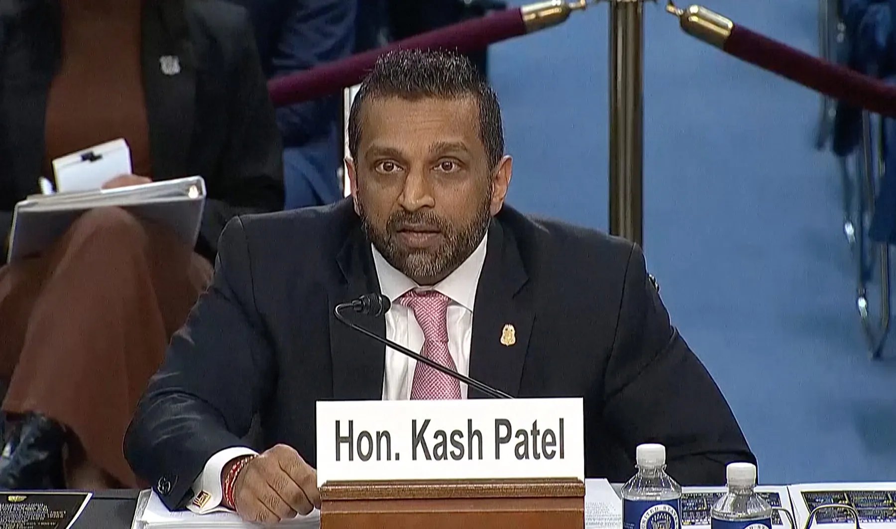 Man in suit and pink tie speaks at a hearing table with a nameplate reading Hon. Kash Patel