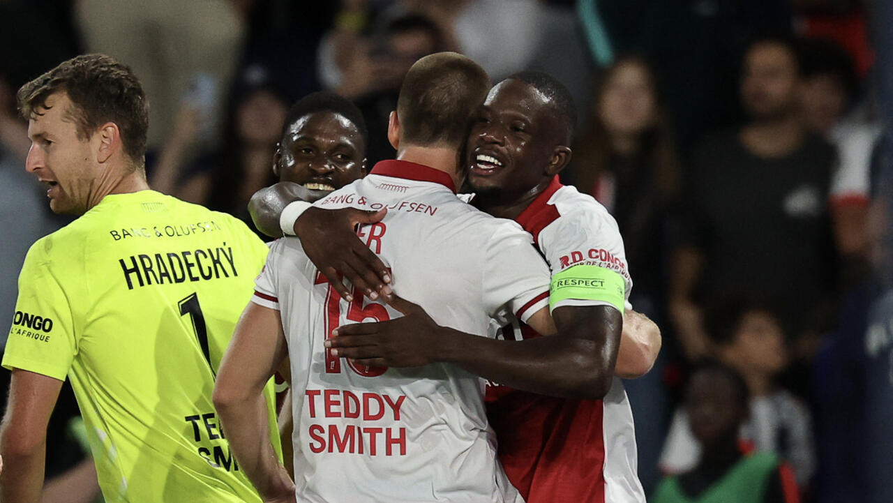 Football players in white and red kits embrace beside a goalkeeper wearing a bright yellow jersey.