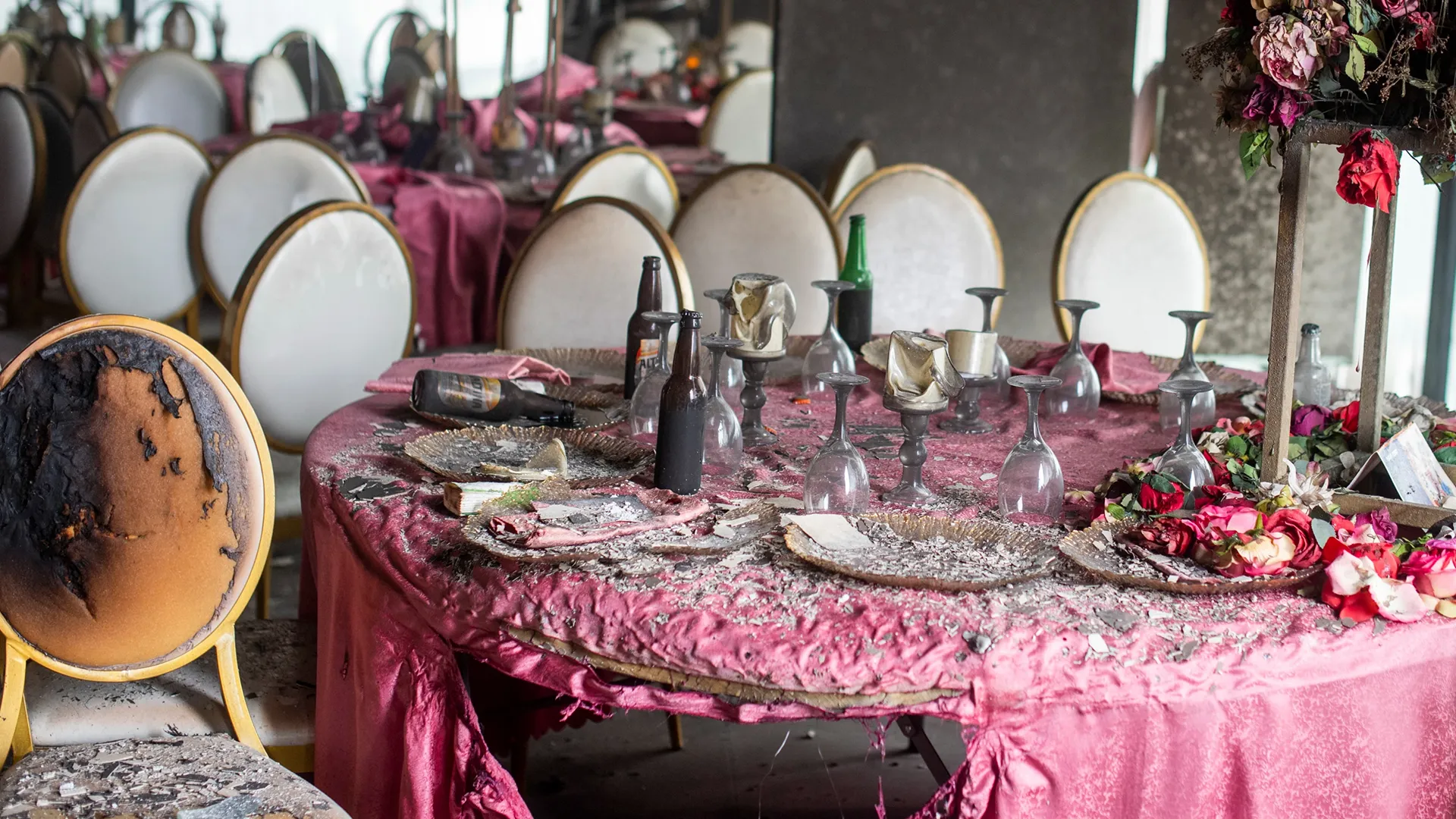 Fire-damaged banquet hall with shattered tableware, bottles, floral decorations and a burned chair.