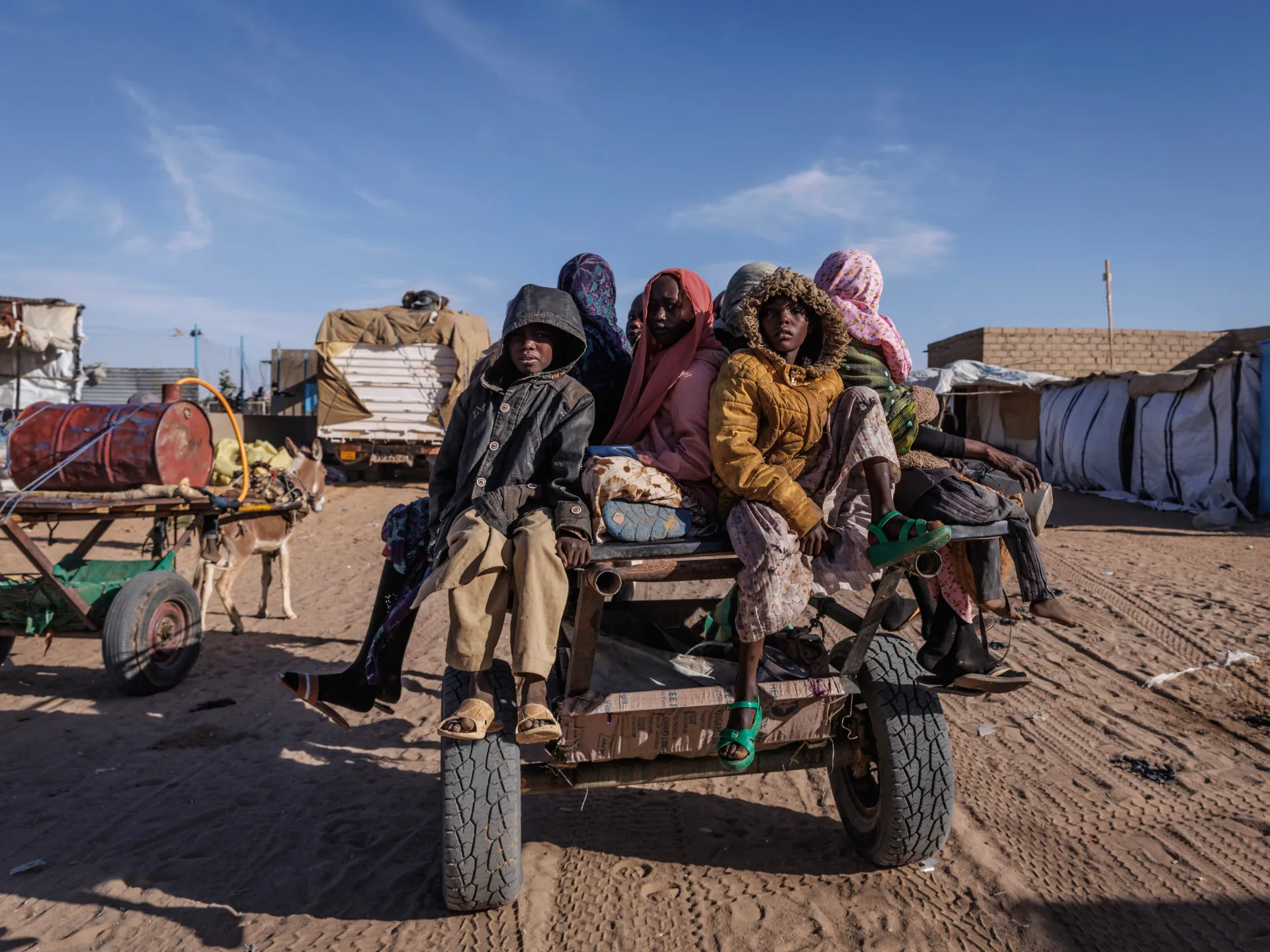 Several people in winter clothing sit on a vehicle amid a dusty settlement under a clear sky.