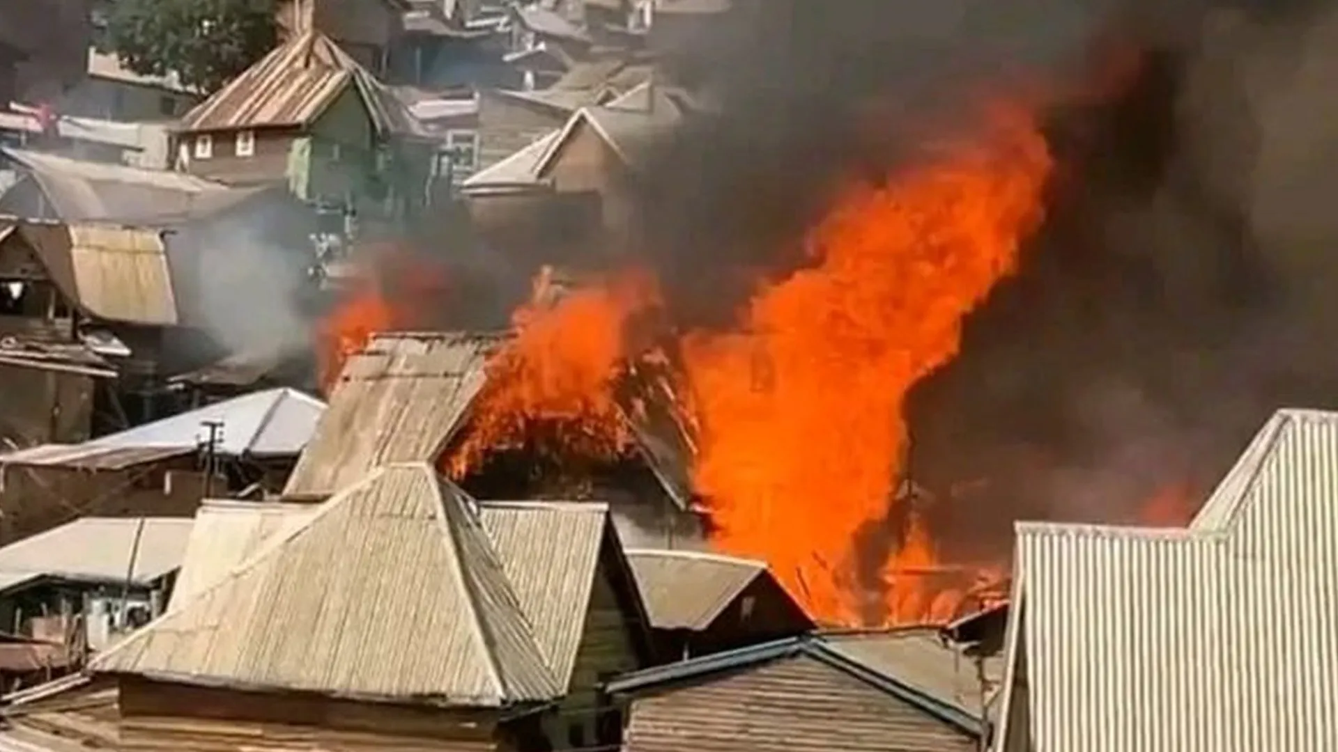 Large flames and thick black smoke rise above corrugated metal rooftops in a densely built neighborhood.