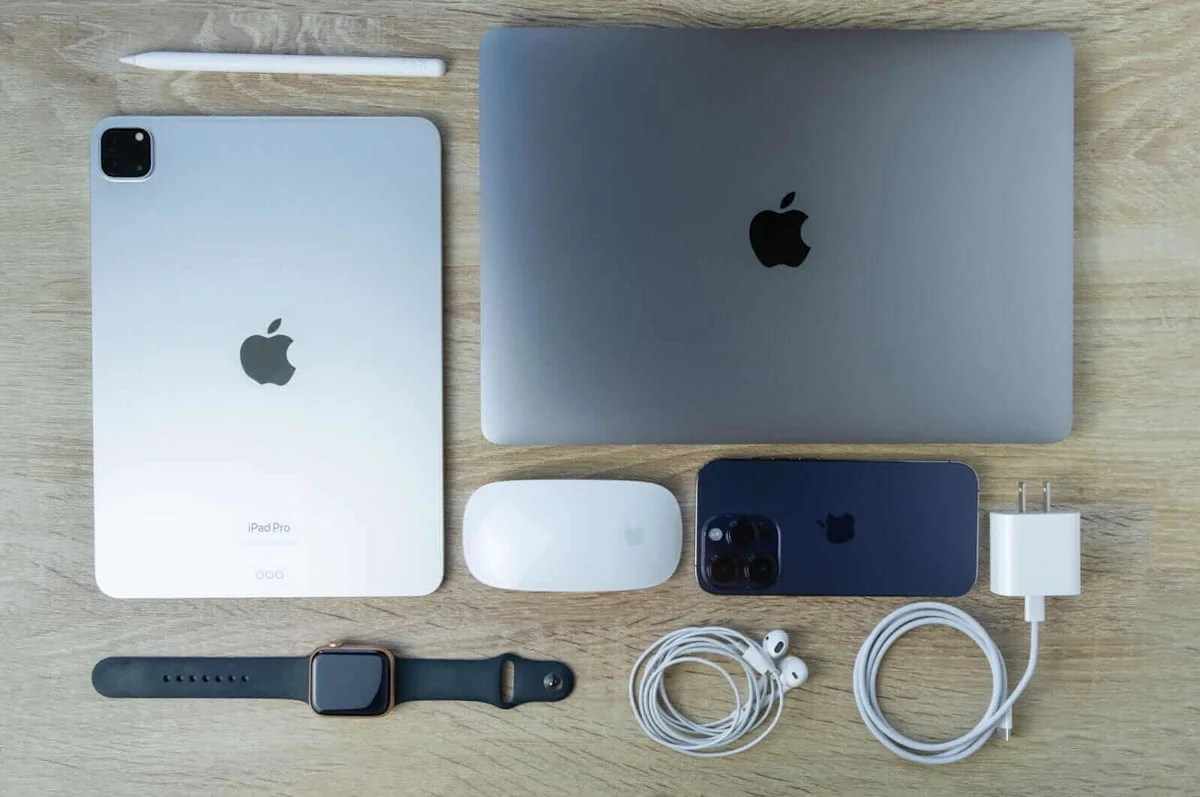 Apple iPad, MacBook, iPhone, Apple Watch, mouse, earbuds, stylus, and charger arranged on a wooden surface.