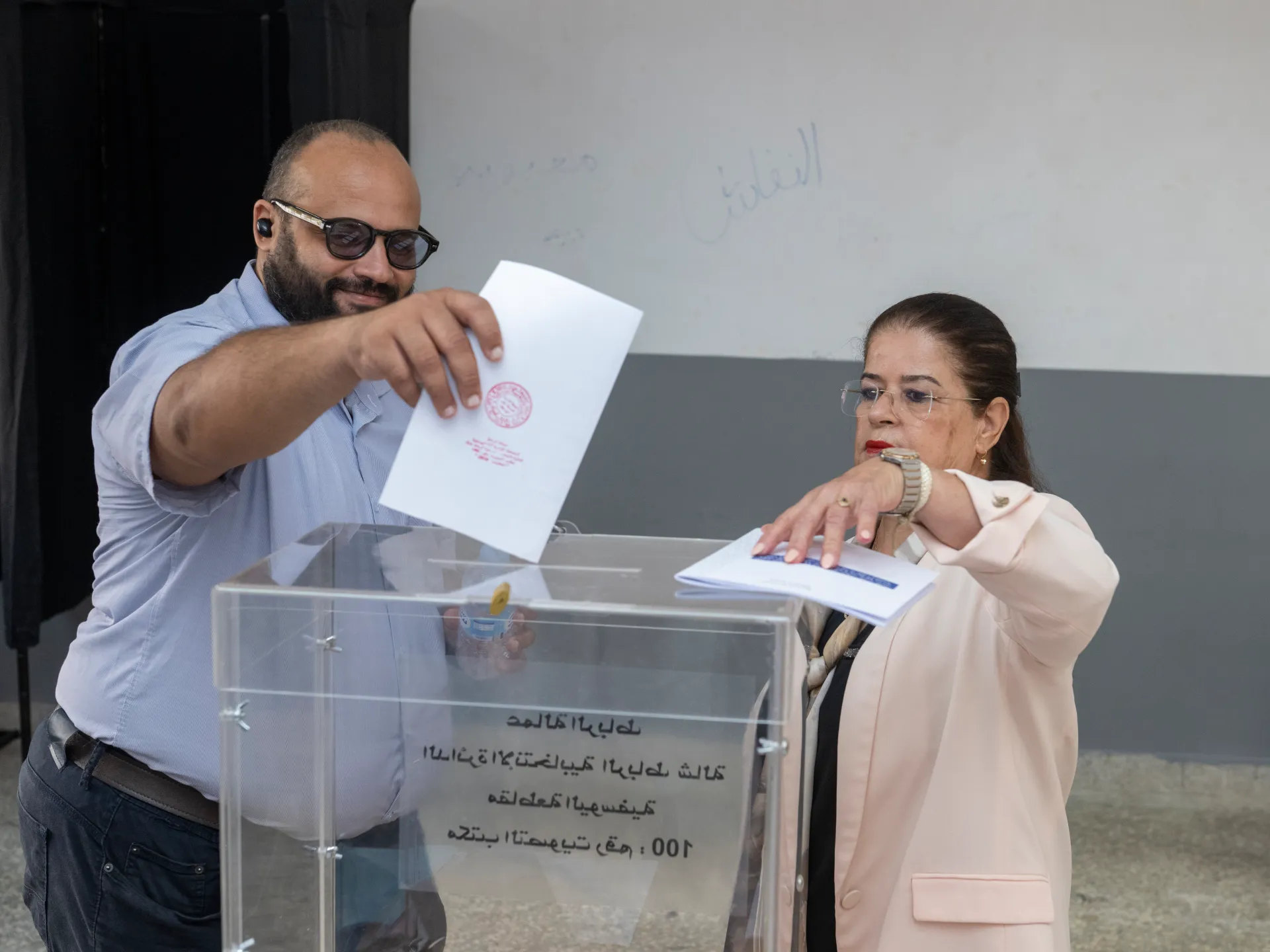 Two election officials handle ballot papers at a transparent ballot box, with Arabic text visible on its front.
