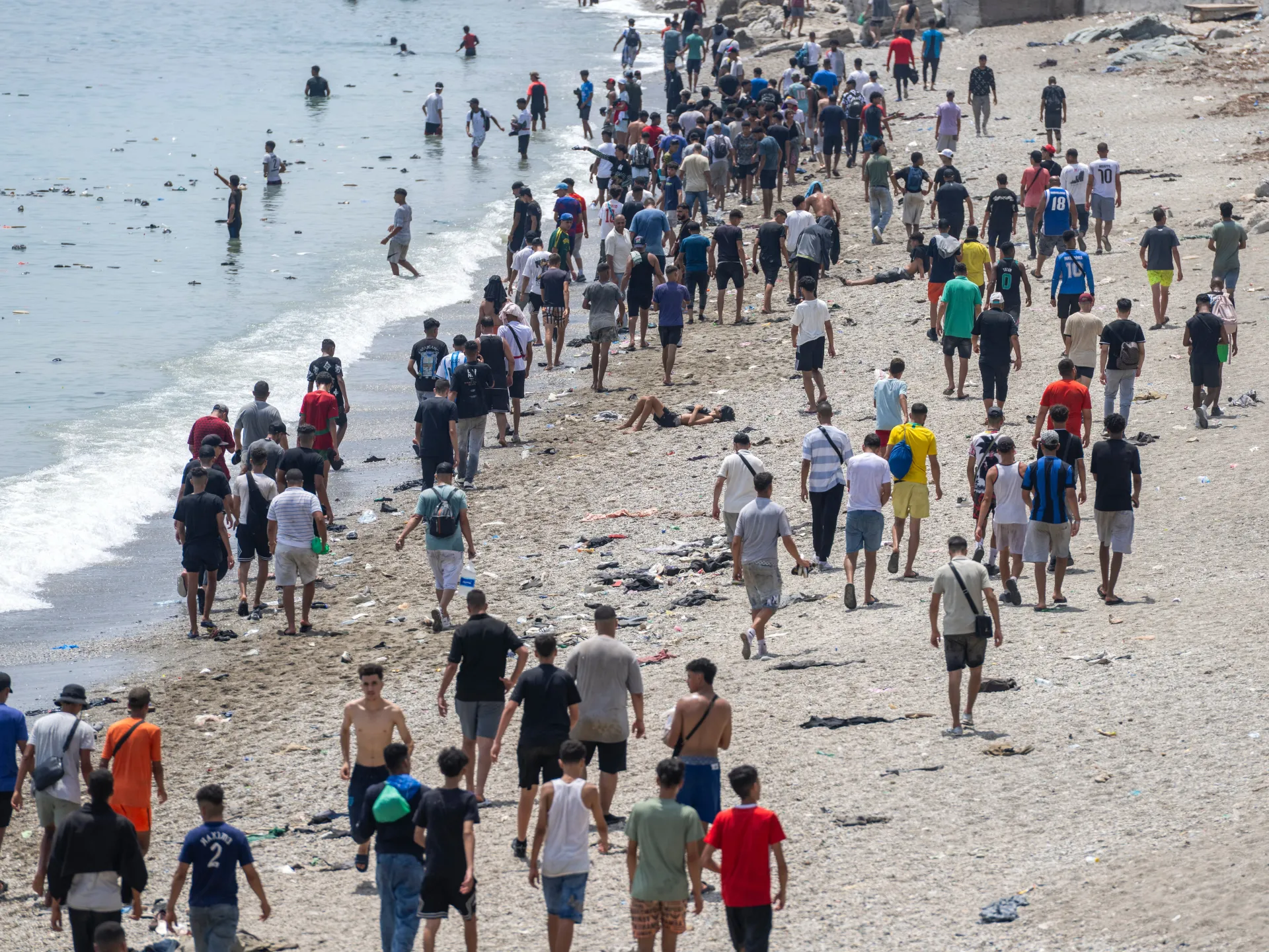 Crowd of people walking along a litter-strewn beach, with others standing in shallow water.
