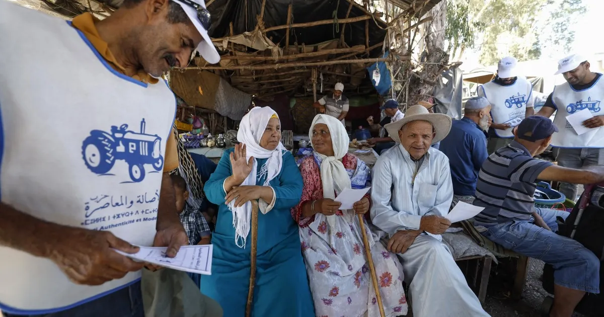 People gather under a rustic shelter, holding papers and wearing matching tractor-logo shirts.