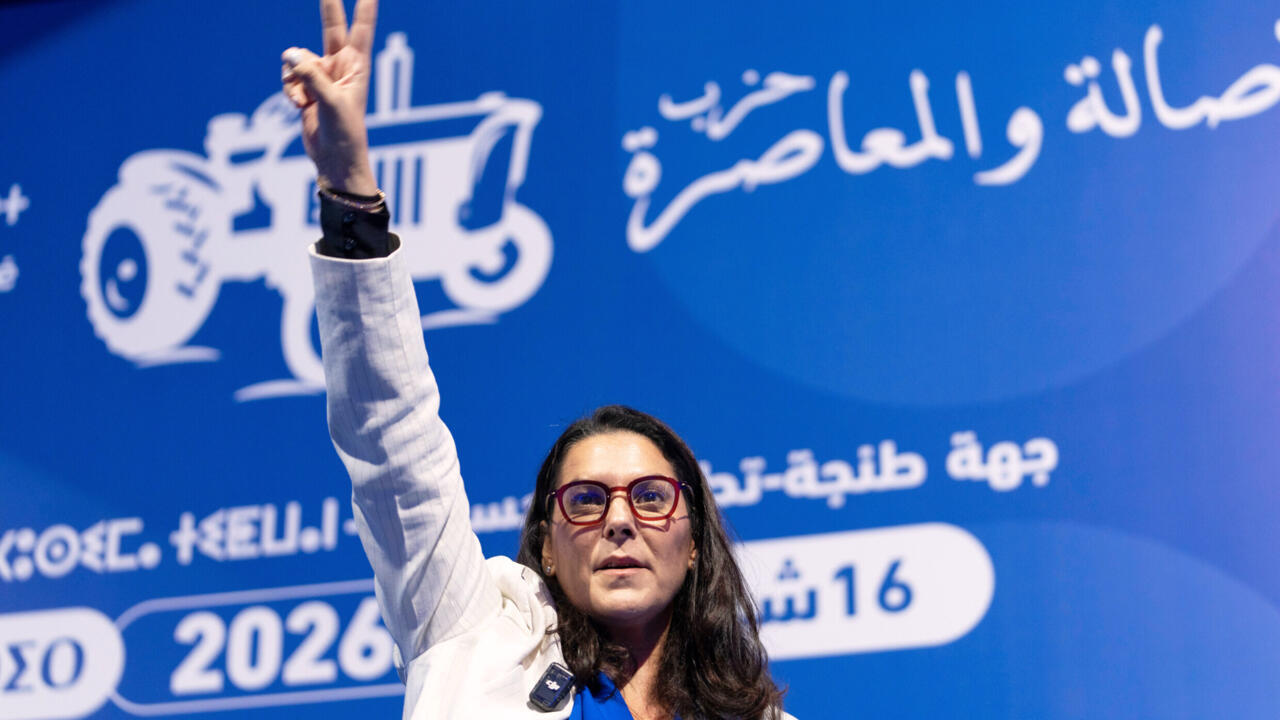 Woman in red glasses and white jacket raises two fingers against a blue Arabic-language backdrop.