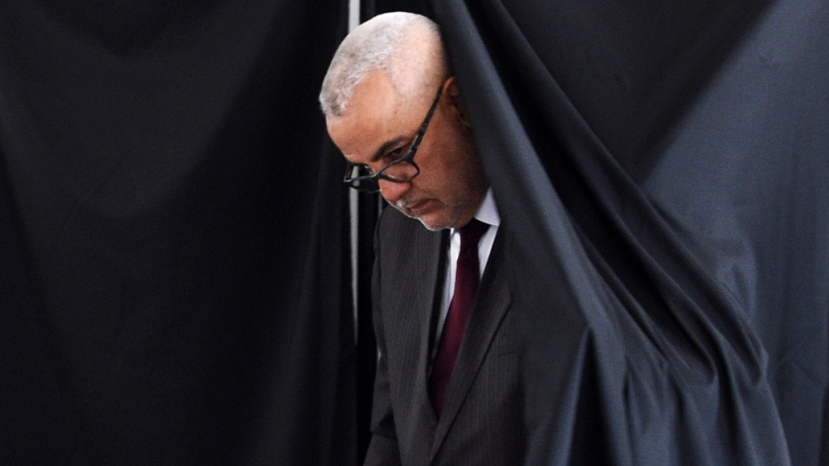 Gray-haired man in glasses and suit walks beneath a dark draped curtain.