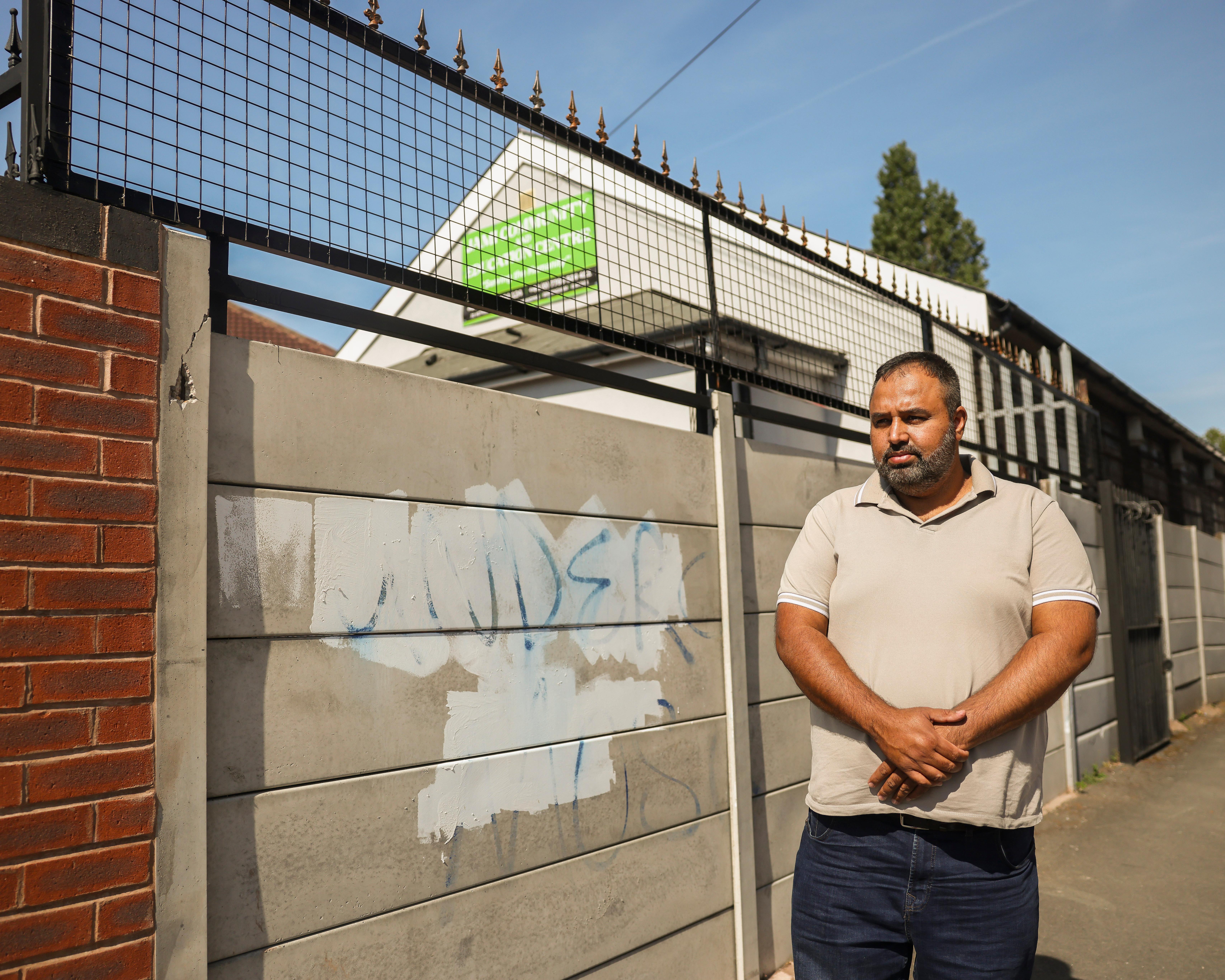 A man stands with folded hands beside a tall metal gate outside a building.