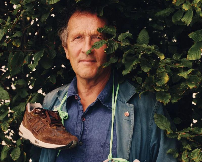 Man standing among leafy branches, holding a brown hiking shoe and wearing a blue shirt.