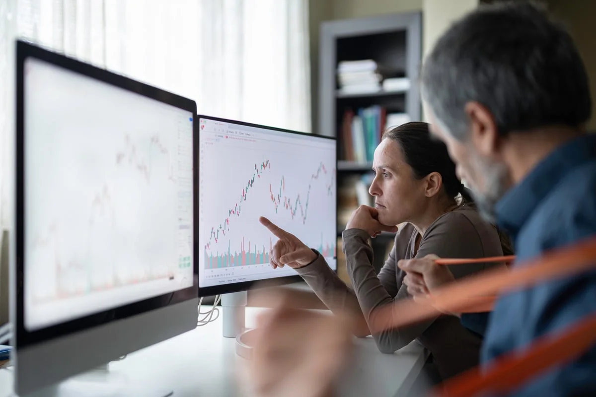 Two colleagues review stock market charts on computer monitors in an office.