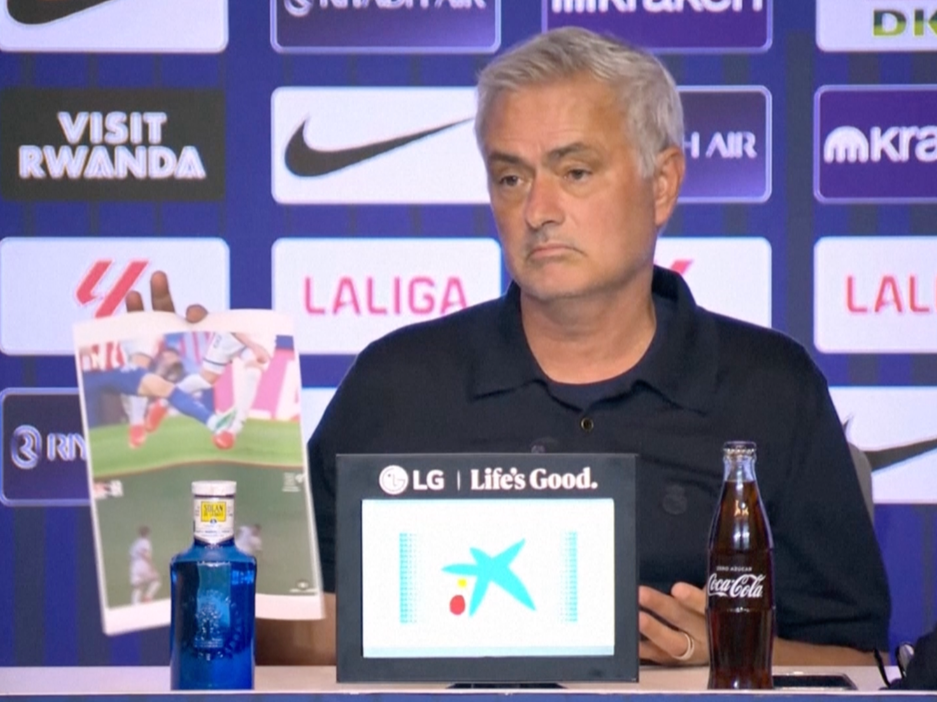 Football manager at a press conference holding a printed match image beside sponsor logos and beverage bottles