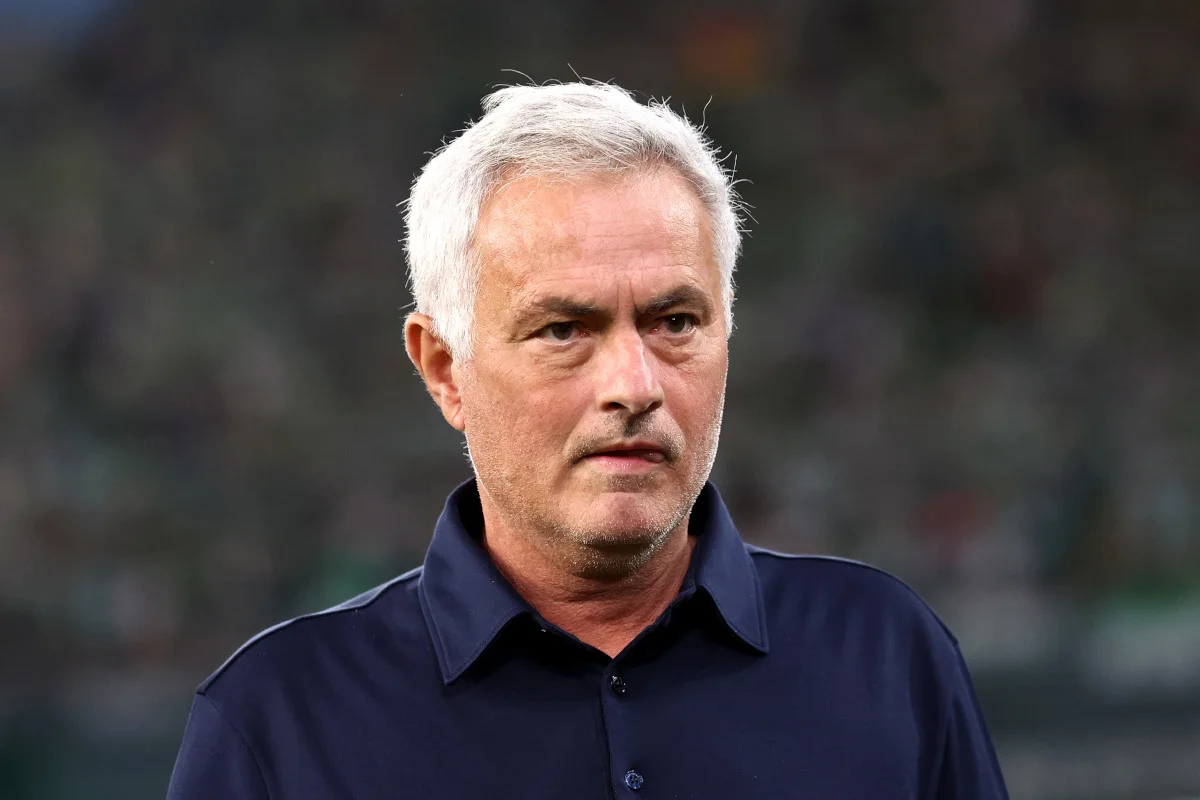 White-haired man in a dark collared shirt looking ahead against a blurred stadium background