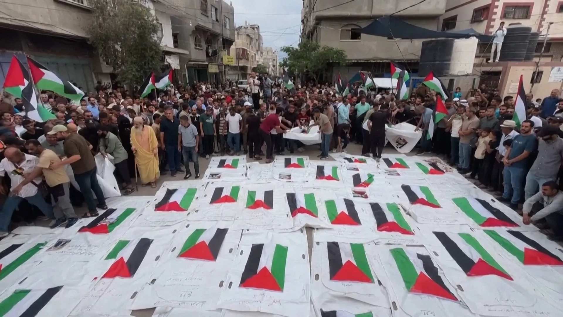 Crowd gathers around white-wrapped bodies marked with Palestinian flags on a crowded street.