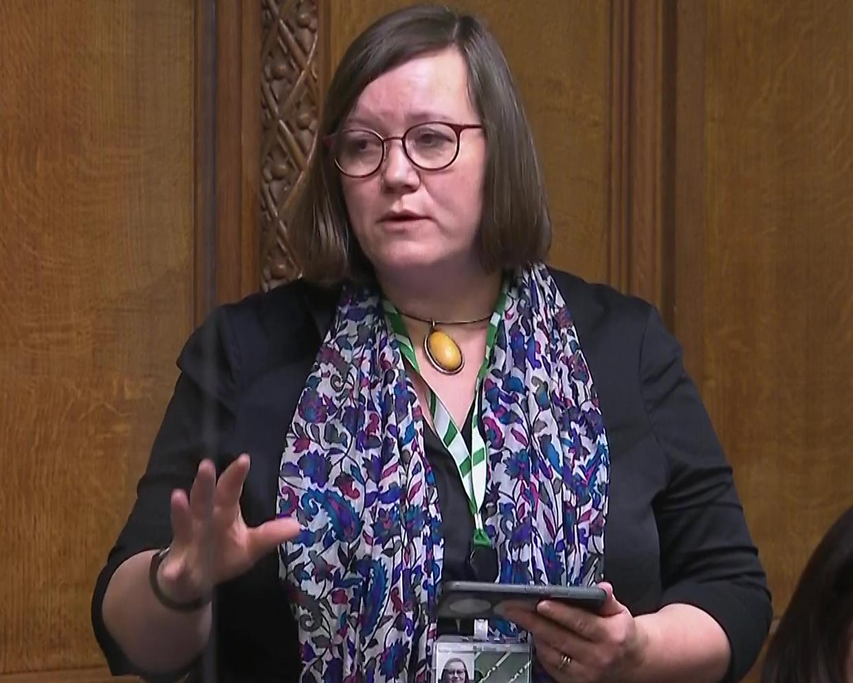 Woman wearing glasses and patterned scarf speaks while holding a tablet in a wood-panelled room
