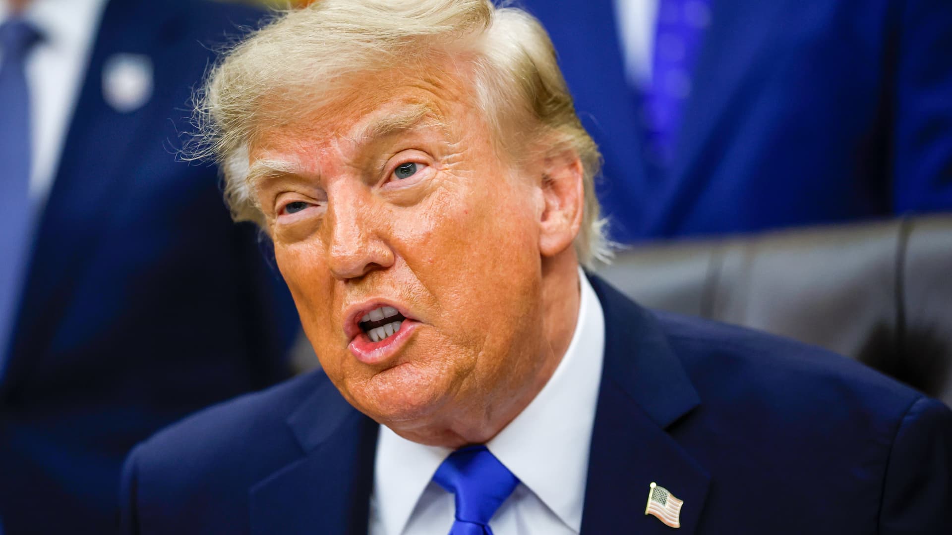 Close-up of Donald Trump speaking in a dark suit and blue tie against a blue backdrop