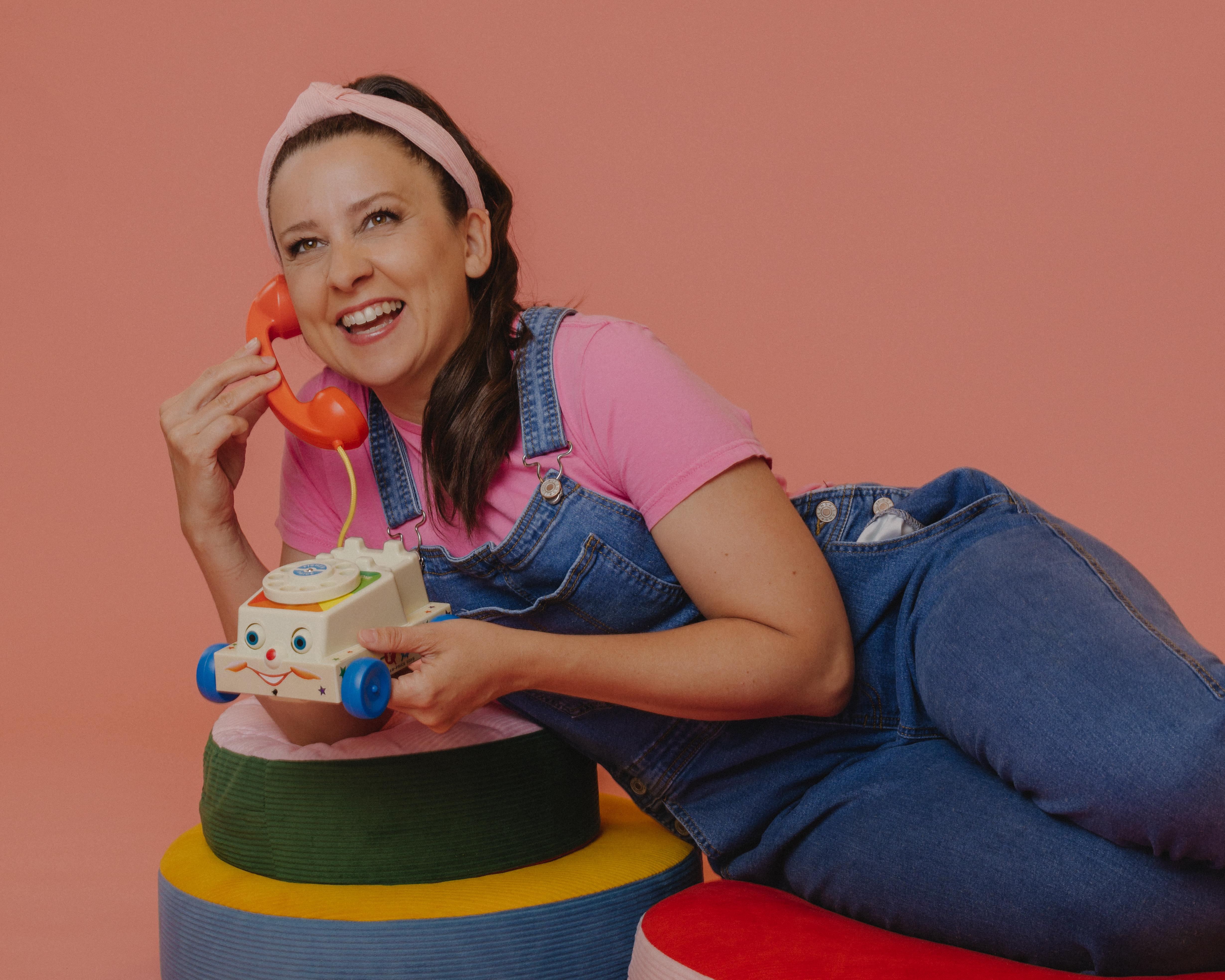 Smiling woman in pink shirt and denim overalls holds a toy telephone and push vehicle.