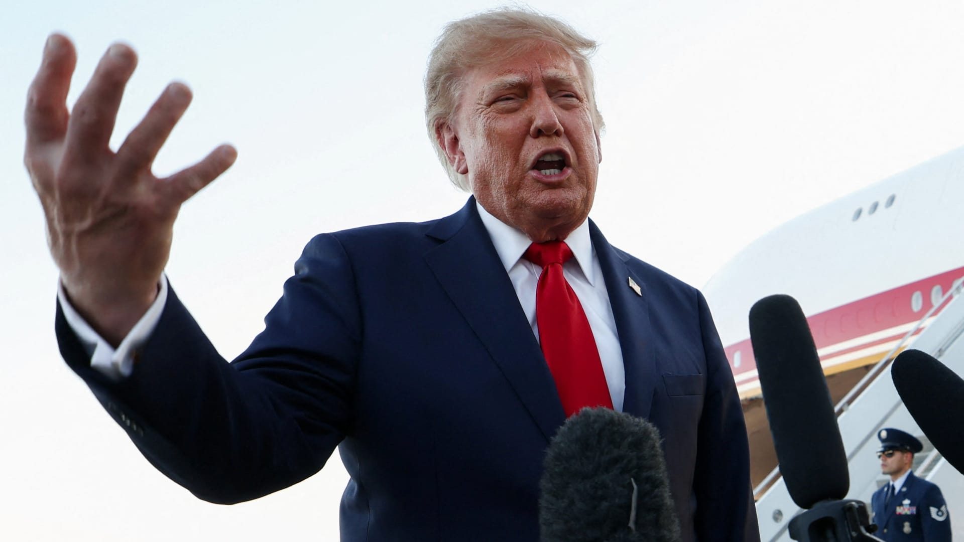Donald Trump gestures while speaking beside microphones near an aircraft staircase.