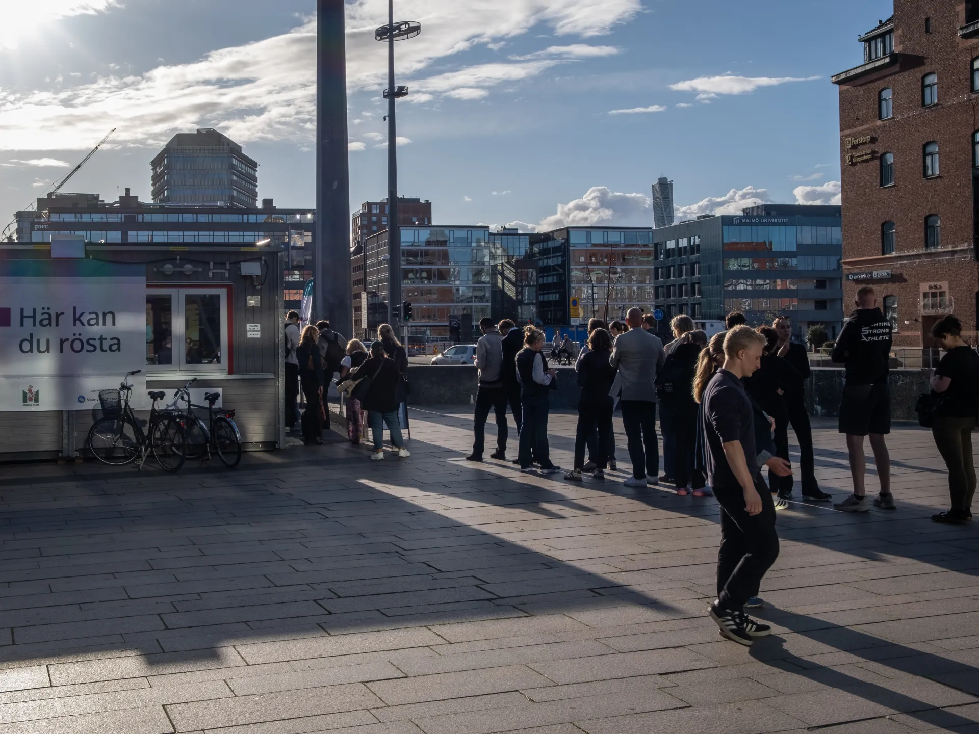 People gather in a Malmö city square beside bicycles, buildings, and a voting booth under sunny skies.