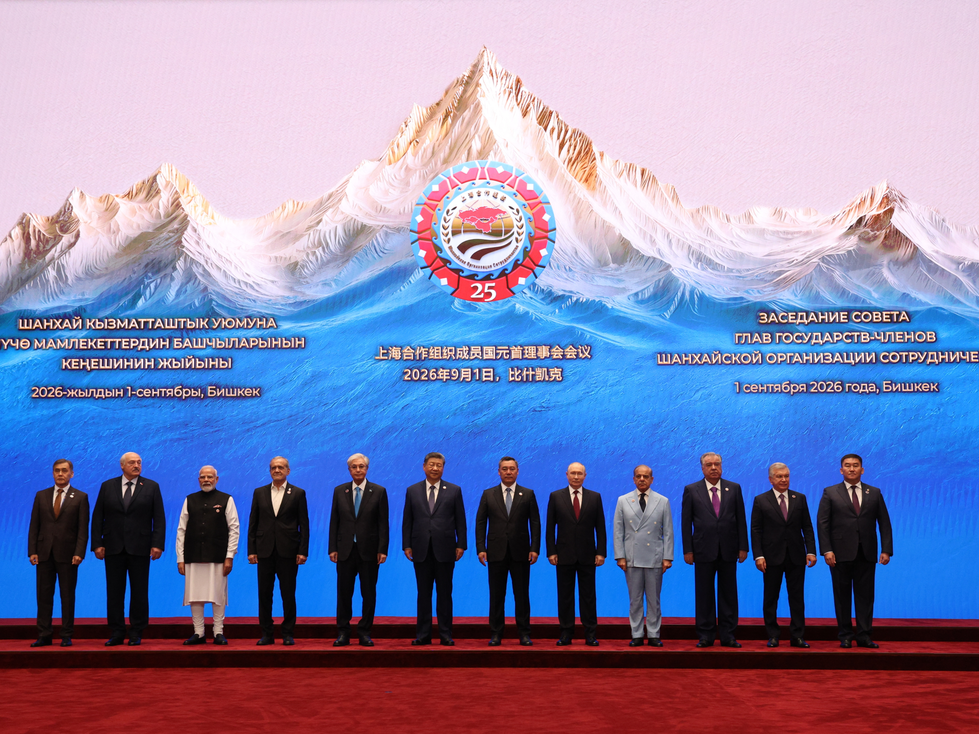 Group of officials pose on a stage beneath a mountain-themed Shanghai Cooperation Organization summit backdrop.