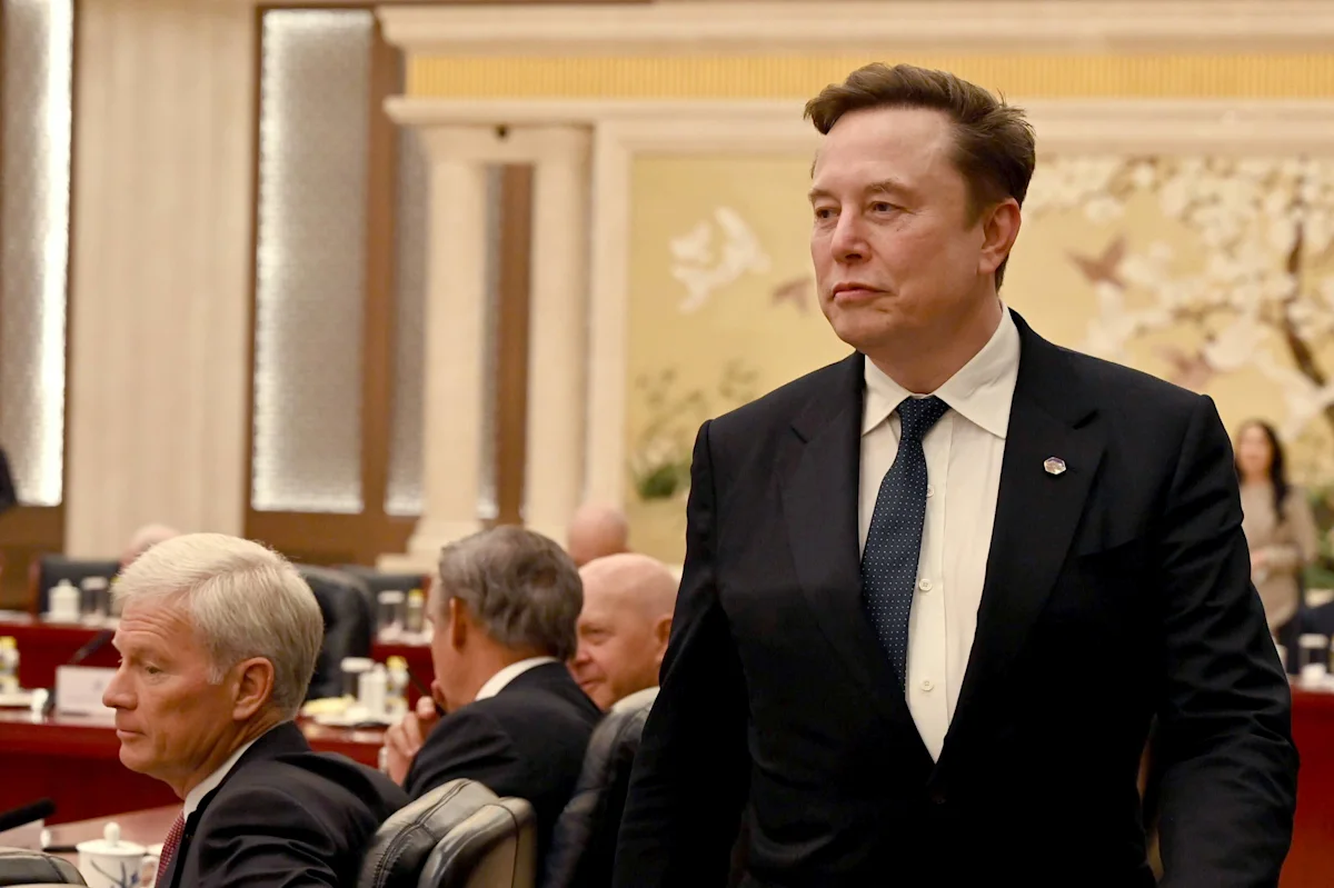Man in black suit and tie standing in a formal meeting room beside seated attendees