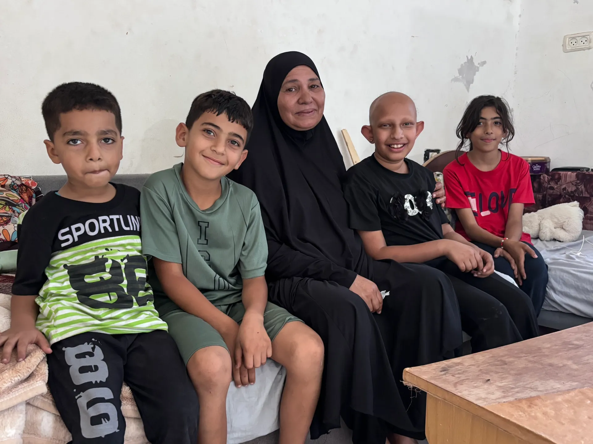 A woman in a black hijab sits with four children on a bed in a modest room.