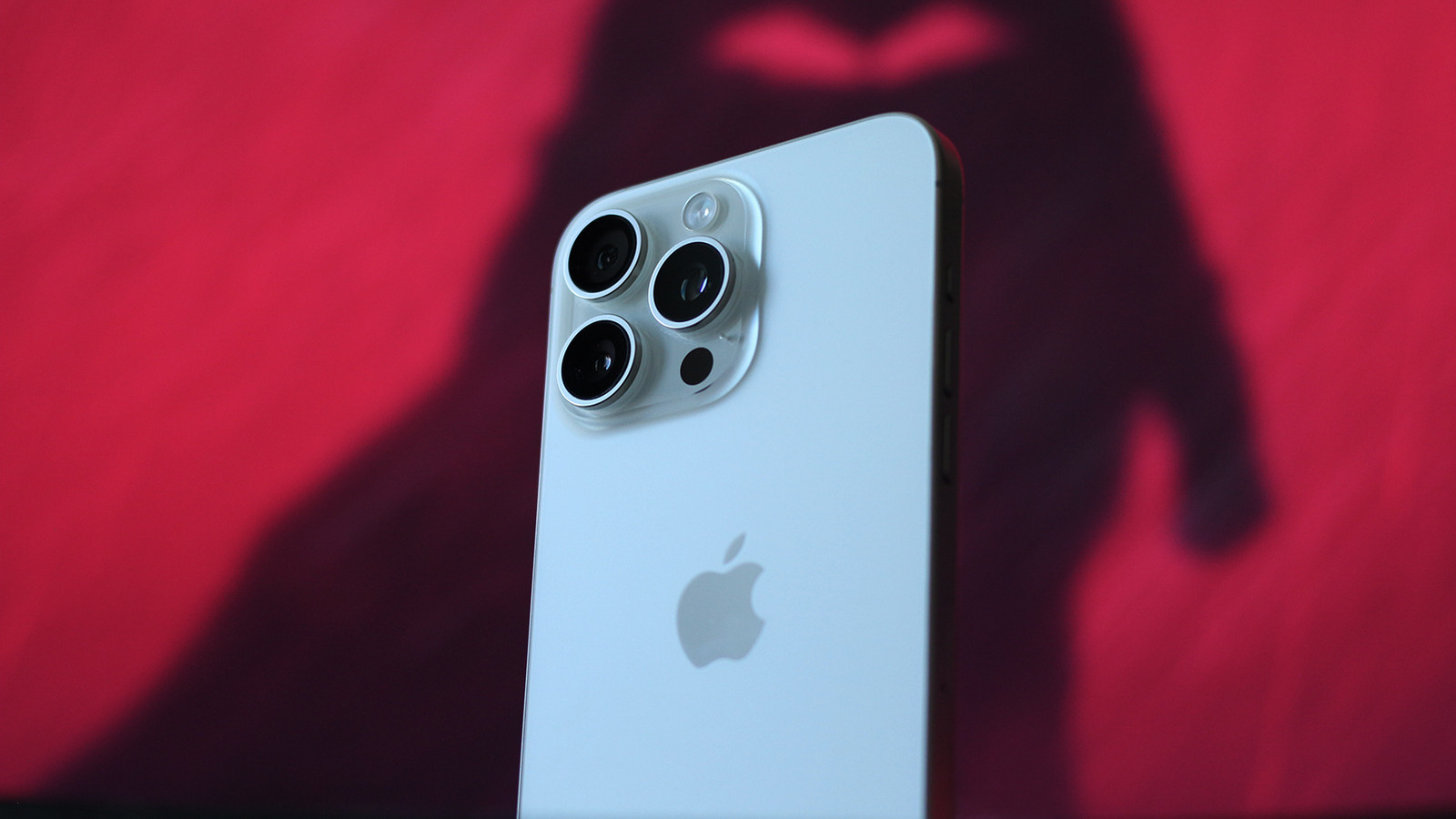A light-colored iPhone viewed from the back, with three camera lenses, against a blurred magenta background.