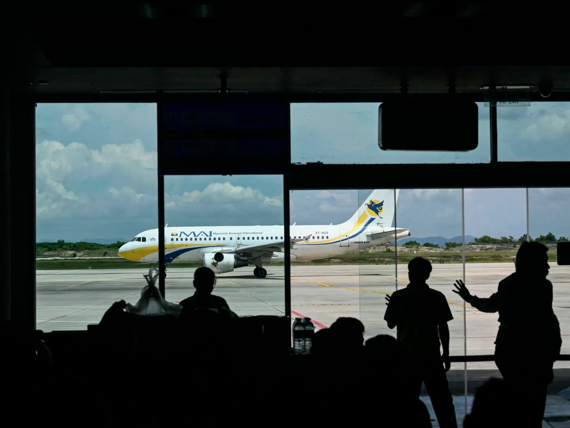 Myanmar Airways International passenger jet viewed through airport terminal windows with silhouetted travelers in foreground
