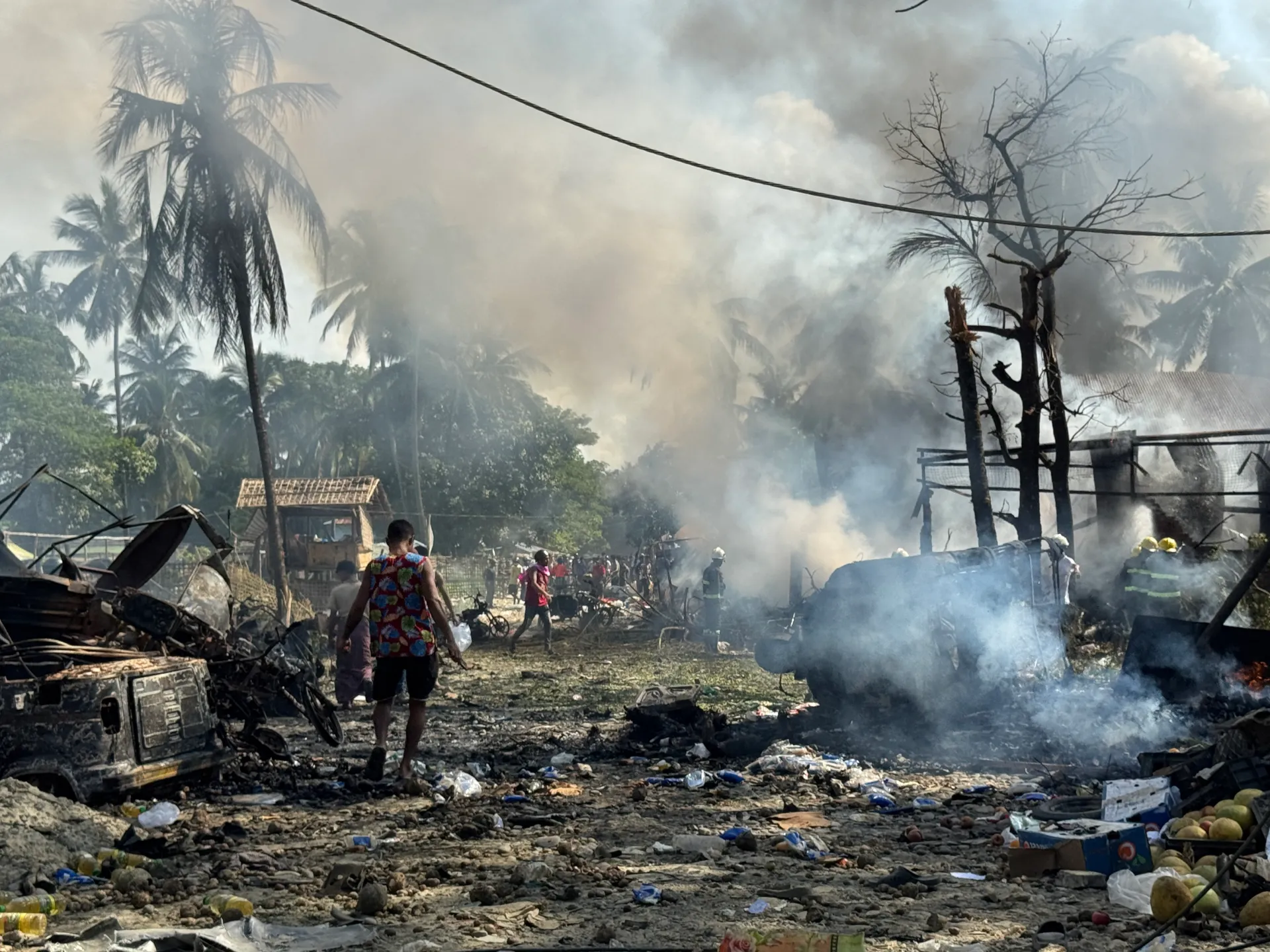 People walk among smoldering wreckage and debris beneath smoke-filled skies, with palm trees and damaged structures nearby.