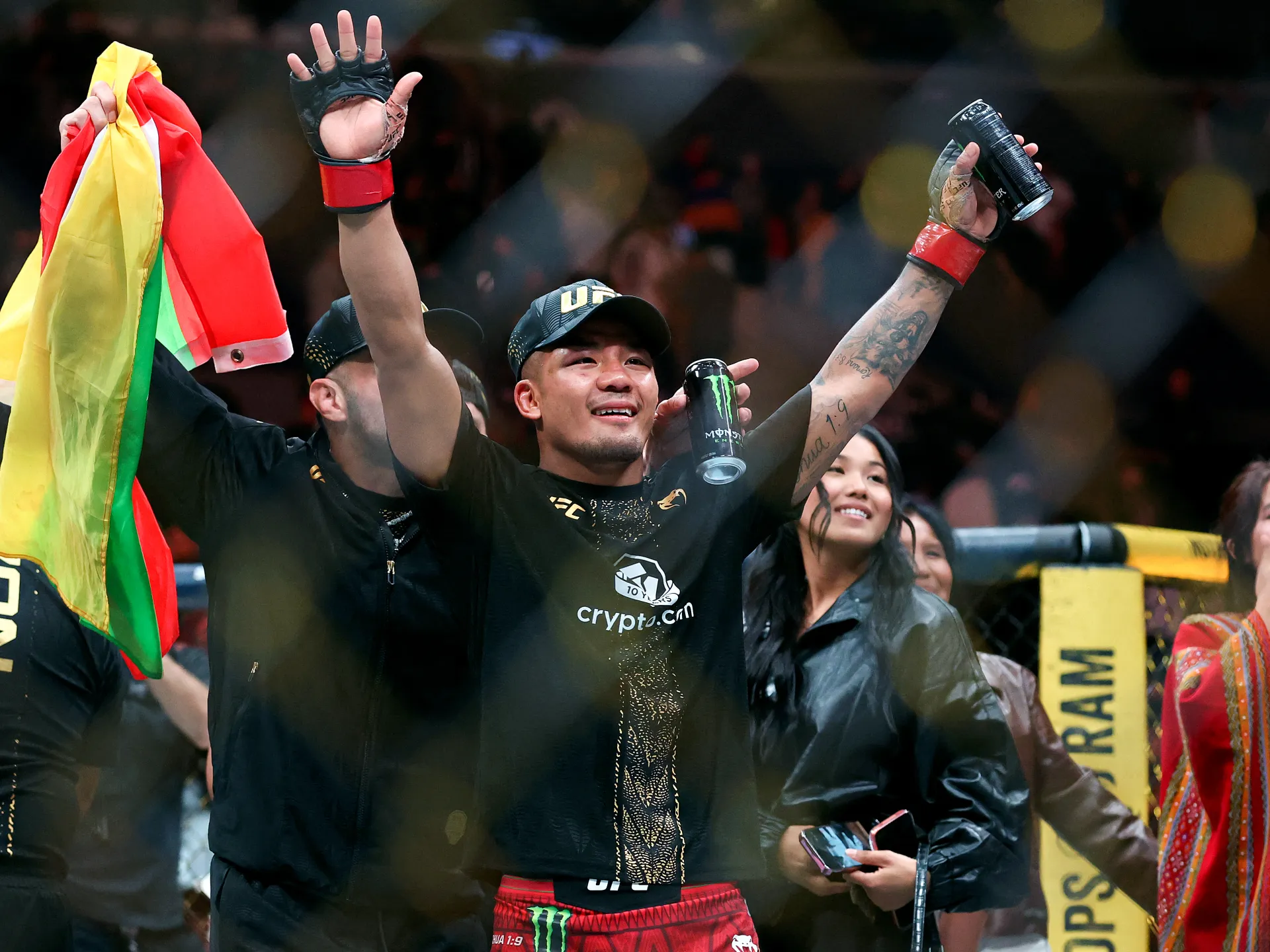 A victorious fighter raises both arms while holding a can, surrounded by teammates and spectators.