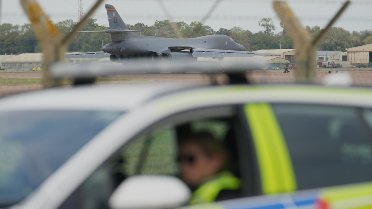 A military bomber sits beyond a blurred police car and fencing at an airbase.