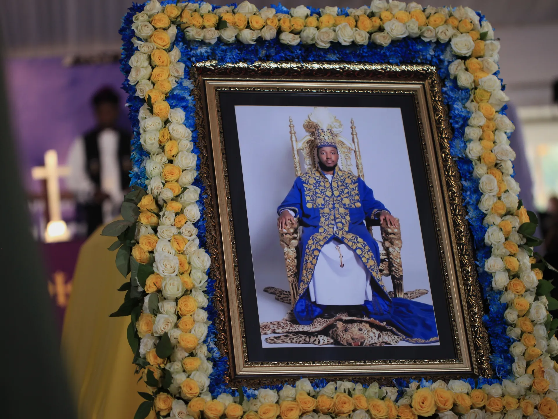 Framed portrait of a man in blue ceremonial clothing surrounded by blue, white and yellow flowers.
