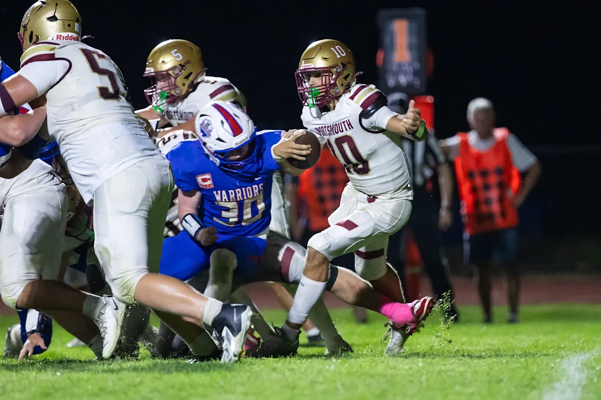 High school football players in white and blue uniforms collide during a nighttime rushing play.