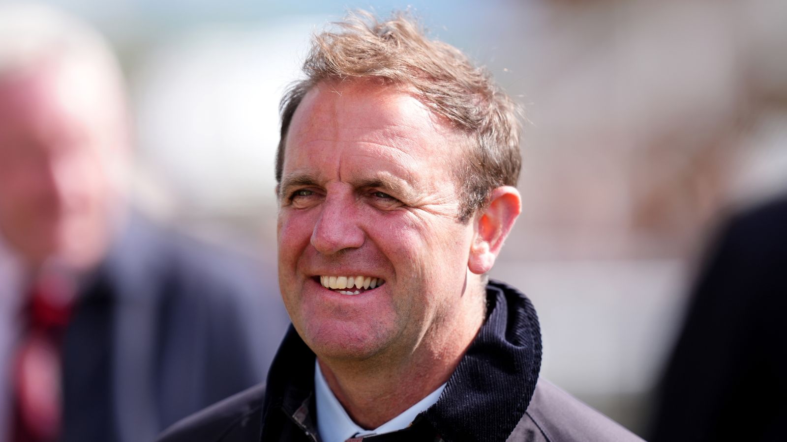 Smiling middle-aged man in a dark coat pictured outdoors with blurred people behind him