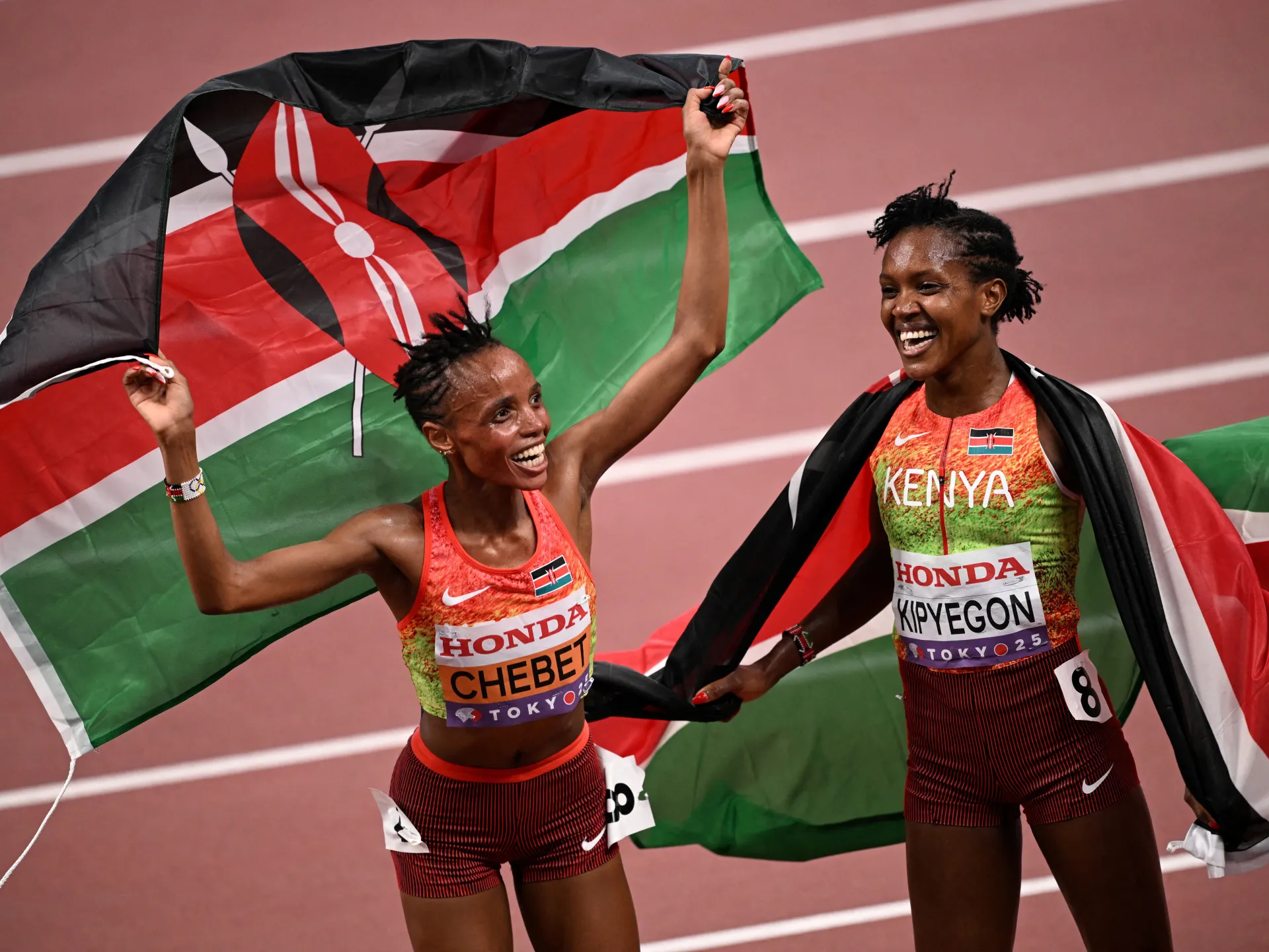 Two Kenyan athletes celebrate while holding a large Kenyan flag on a track.