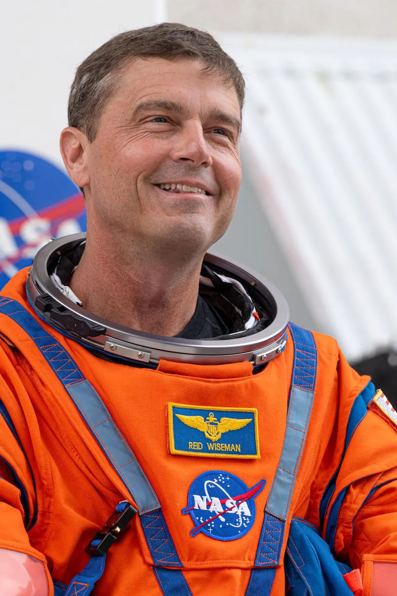 Smiling astronaut wearing an orange NASA spacesuit with blue harness straps and mission patches