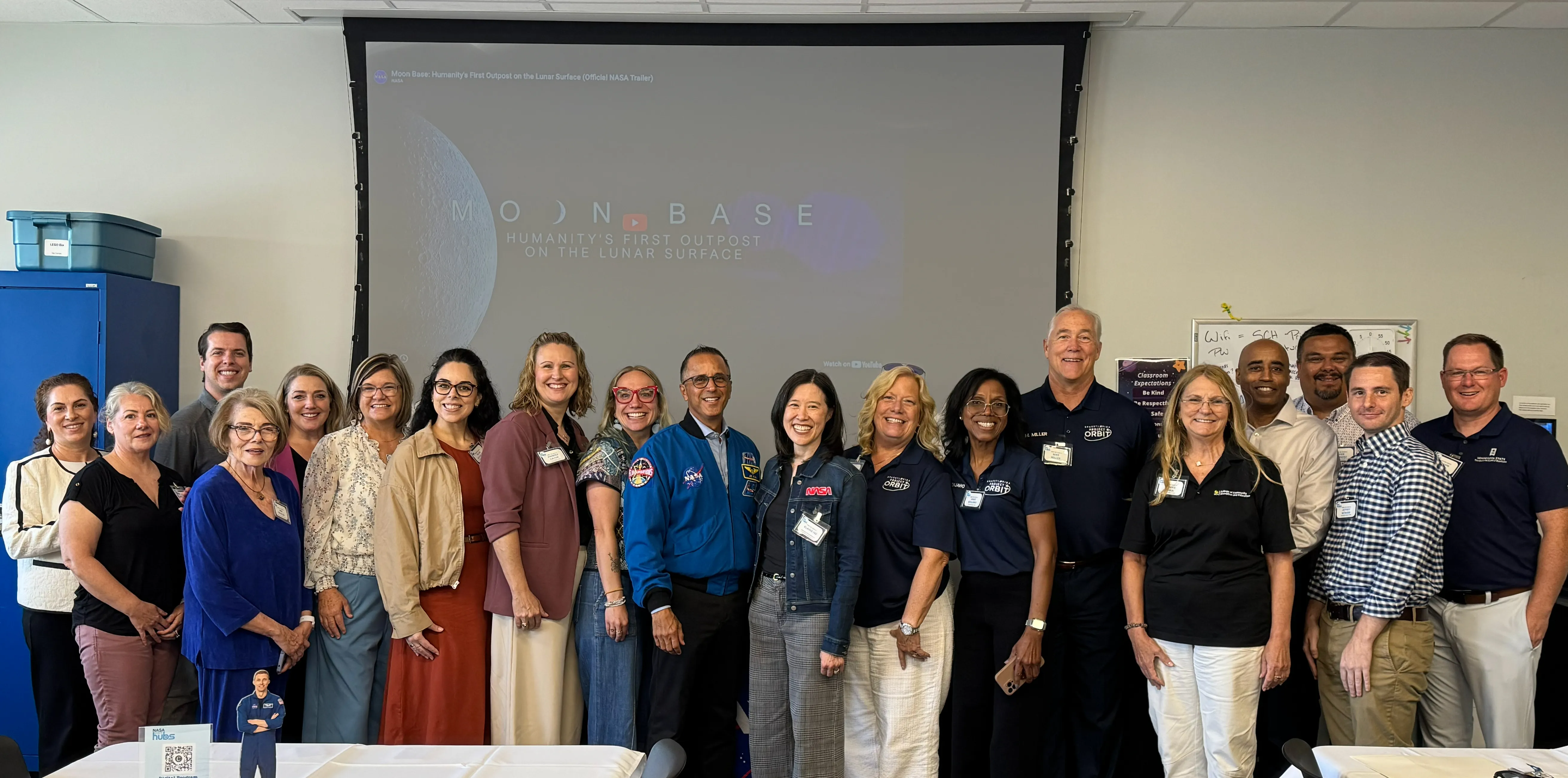 Group of adults posing in a classroom beneath a projected Moon Base presentation.