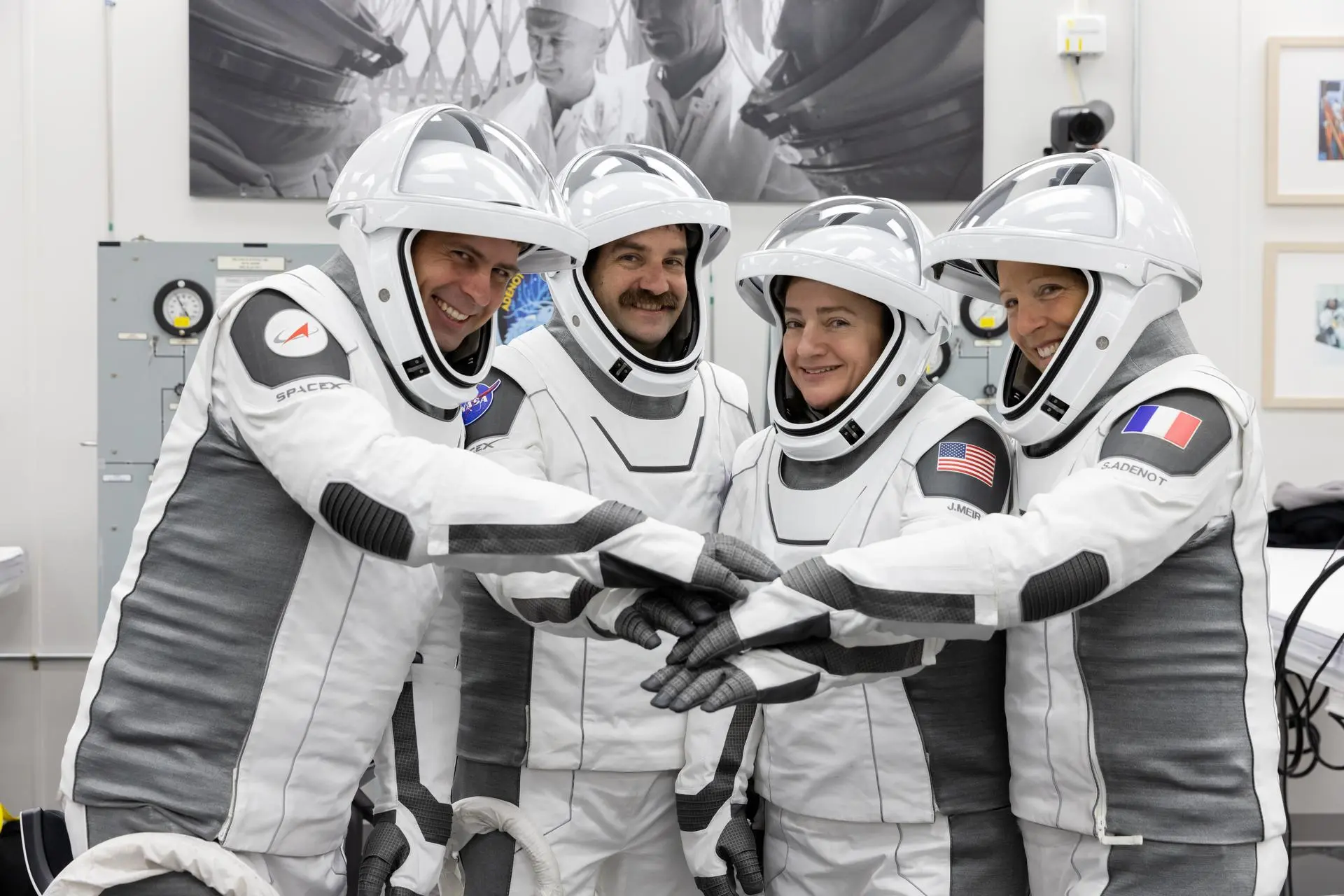 Four astronauts in white spacesuits smile and stack gloved hands during a group photo.