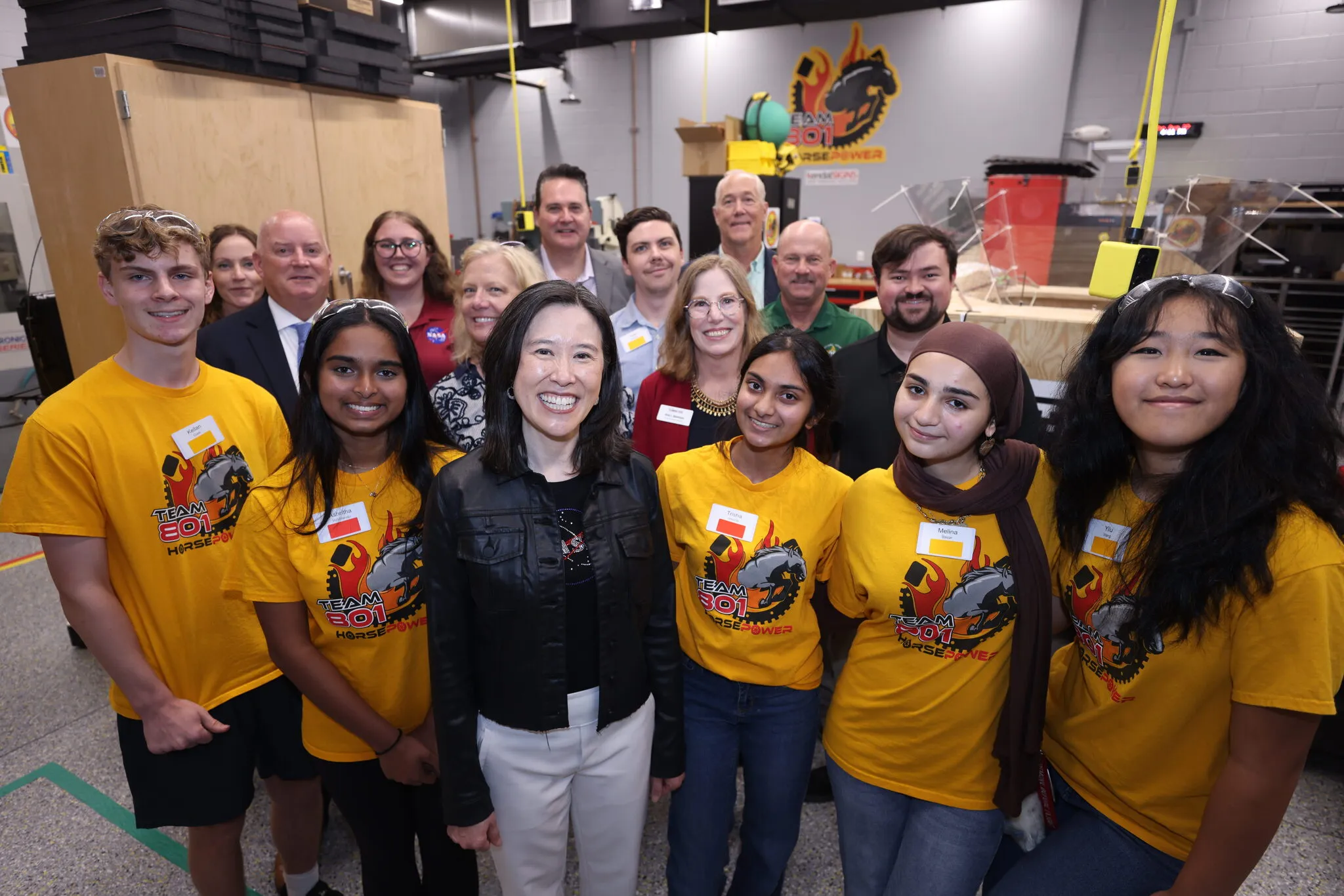 Students and adults pose together in a robotics facility, several wearing yellow Team 301 shirts.