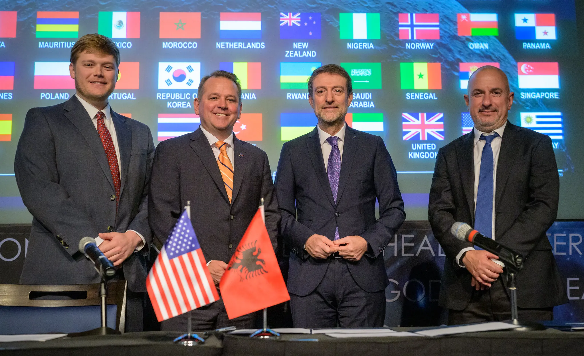 Four officials stand before international flags with U.S. and Albanian flags displayed in front.