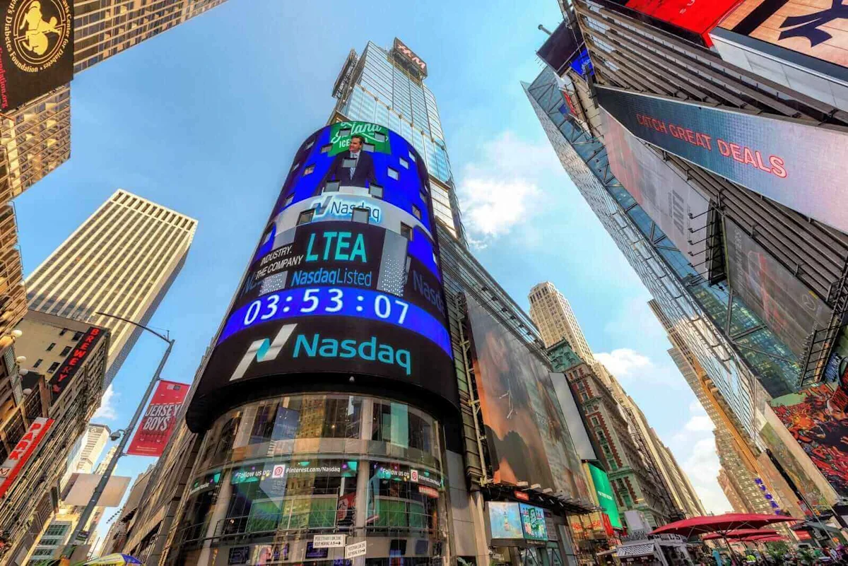 Low-angle view of Times Square buildings with large Nasdaq electronic billboards under a blue sky.