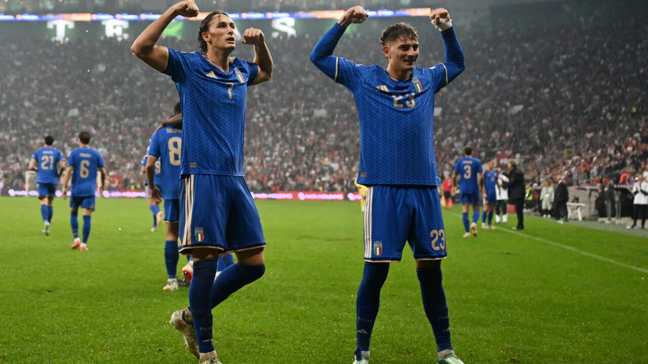 Two Italy players in blue kits raise their arms on the pitch as teammates walk away.