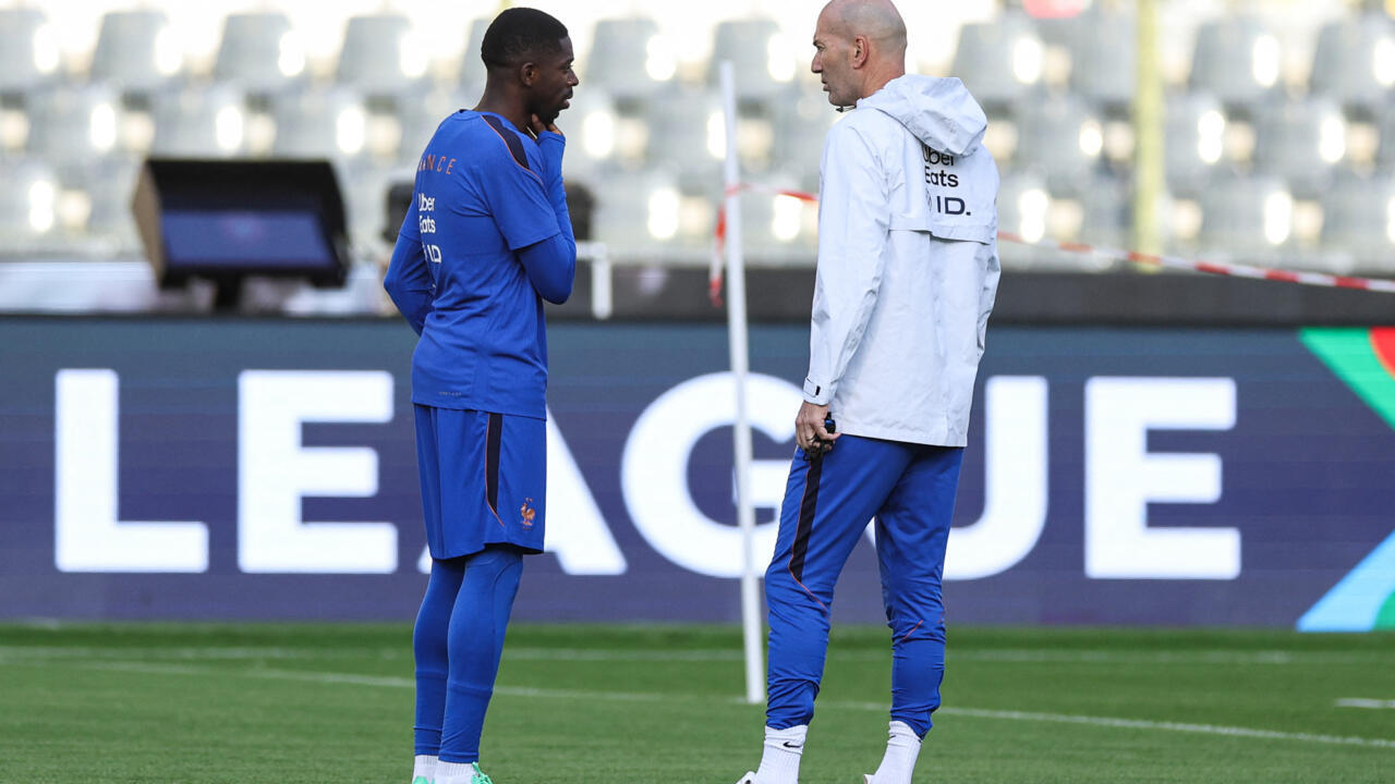A French player in blue speaks with a bald coach in a white jacket on the pitch.