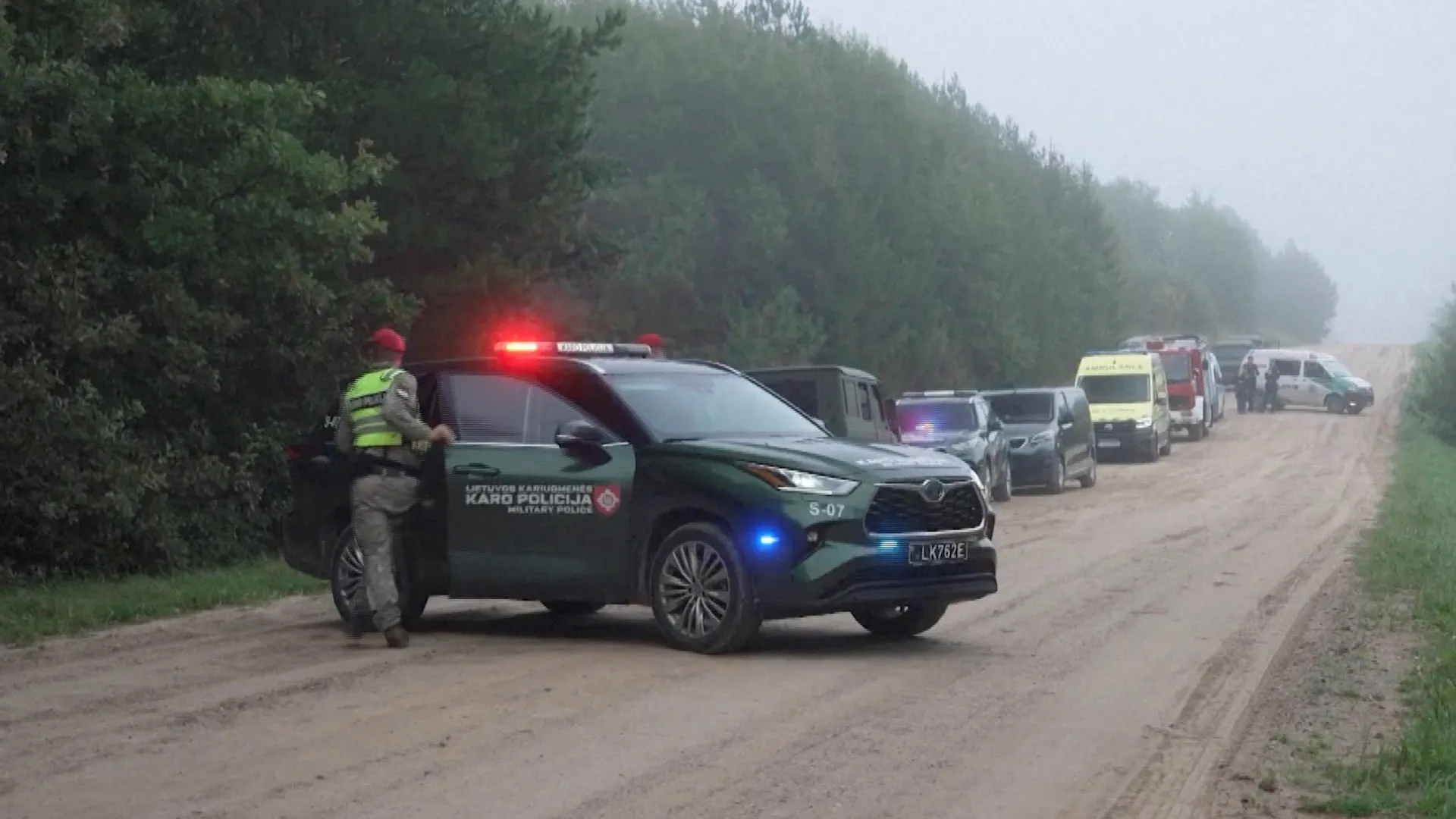Police vehicle and emergency responders block a foggy dirt road lined with trees.