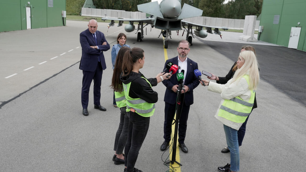 Officials and journalists gather for a media briefing in front of a fighter jet.