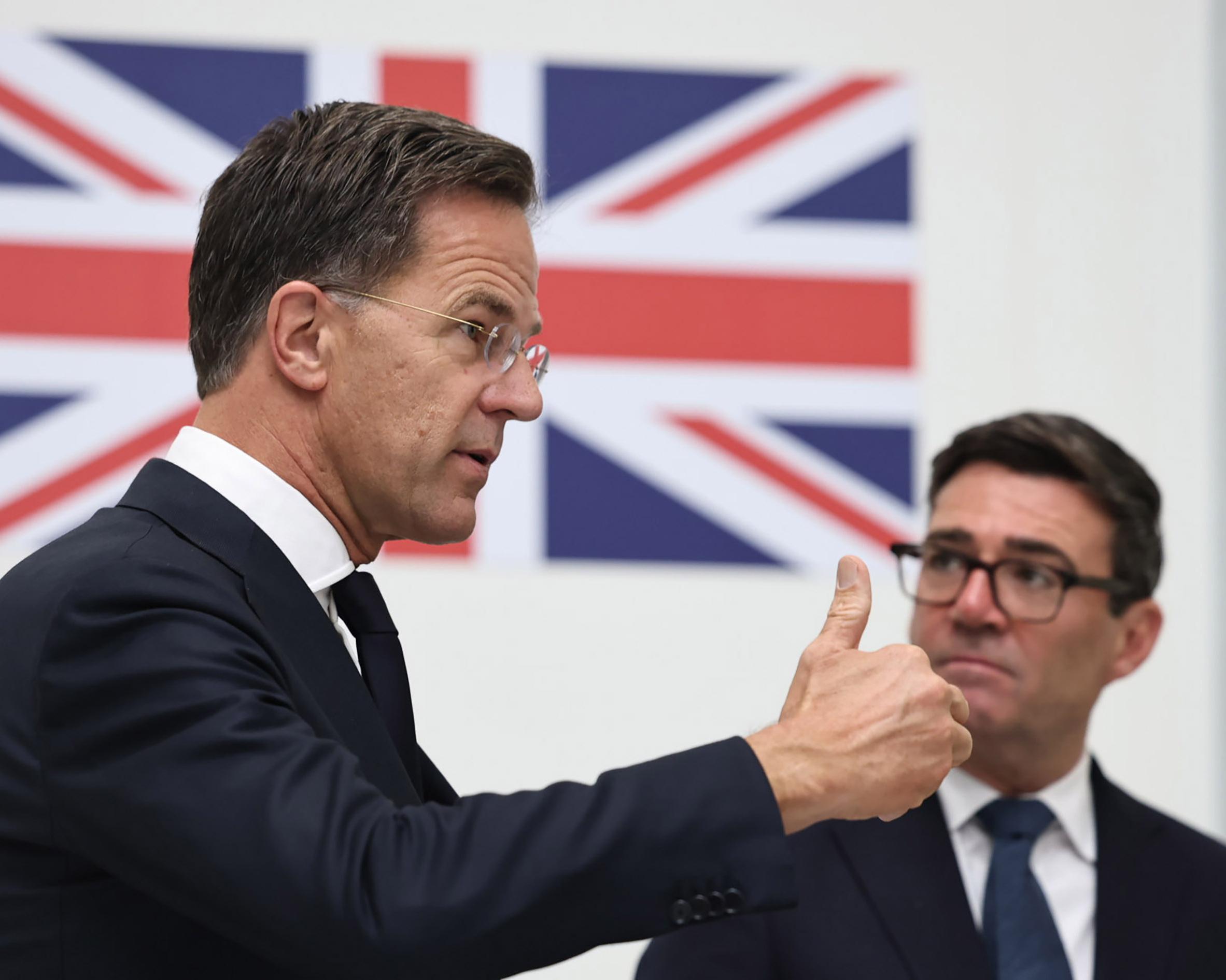 Man in suit gestures in front of Union Jack-style flags as another suited man looks on.