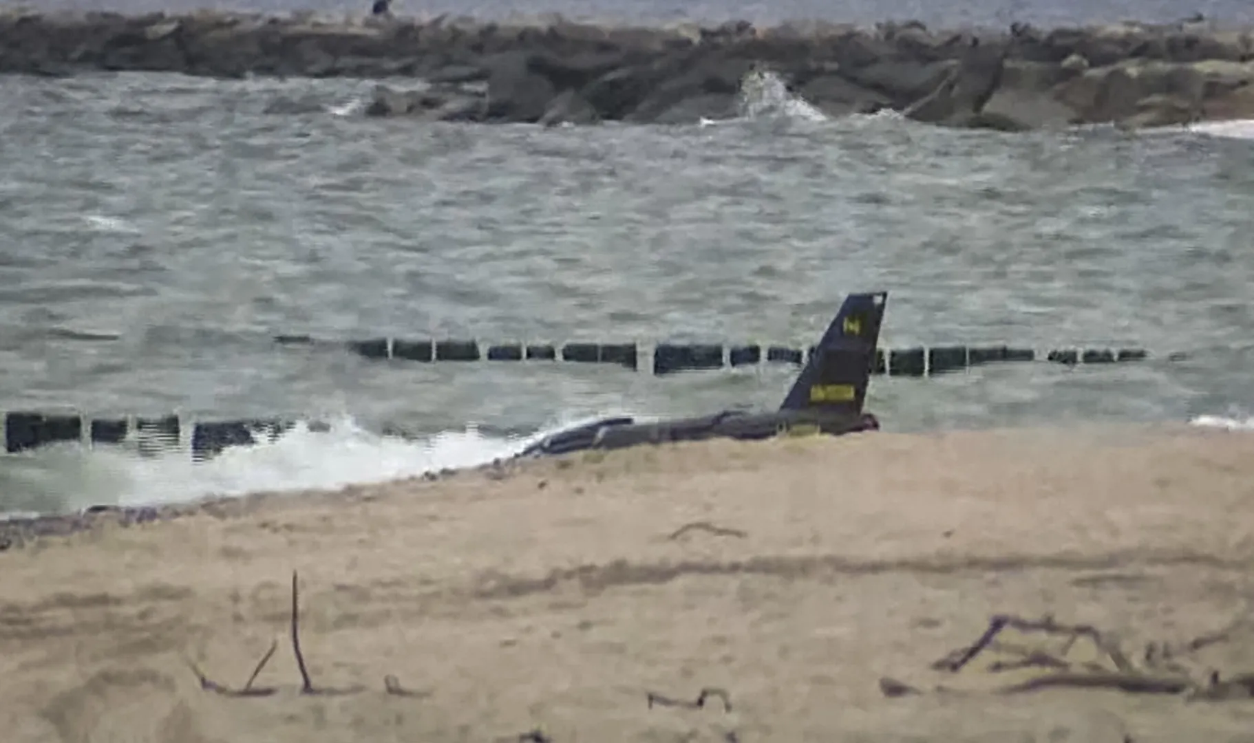 A dark military aircraft tail rests partly submerged near a sandy shoreline and breakwater.