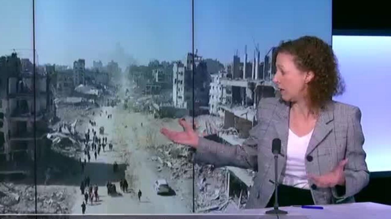 Woman in blazer gestures beside a screen showing heavily damaged buildings and rubble.