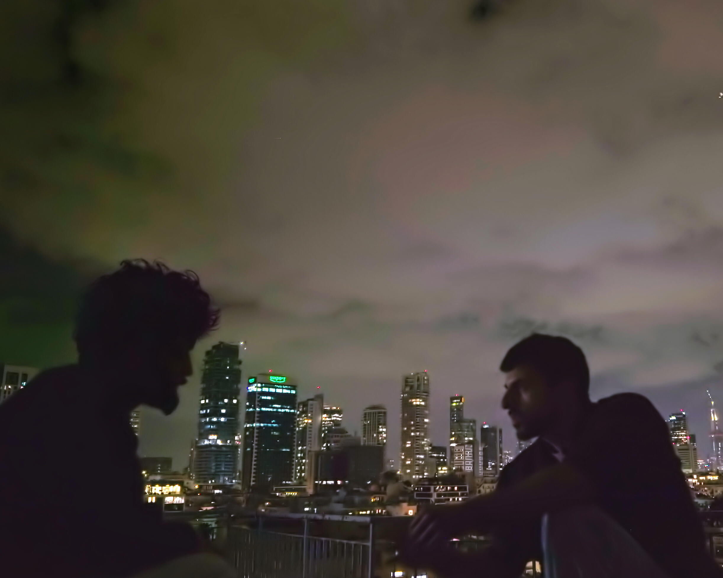 Two silhouetted people stand before a brightly lit city skyline beneath a cloudy night sky.