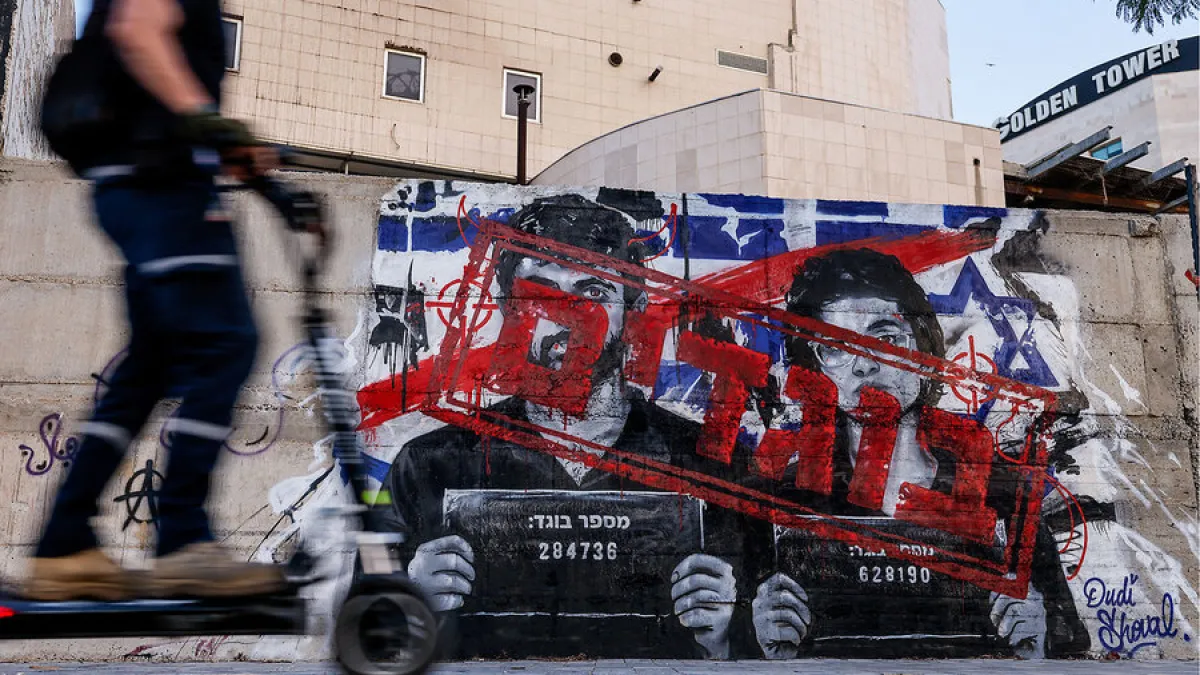 Blurred rider passes a mural depicting two men behind bars with red graffiti and Hebrew text.