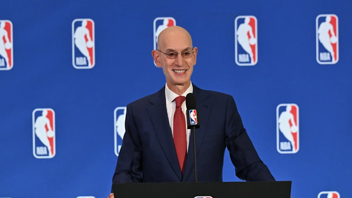 Bald man in suit and red tie speaks at podium before an NBA logo backdrop